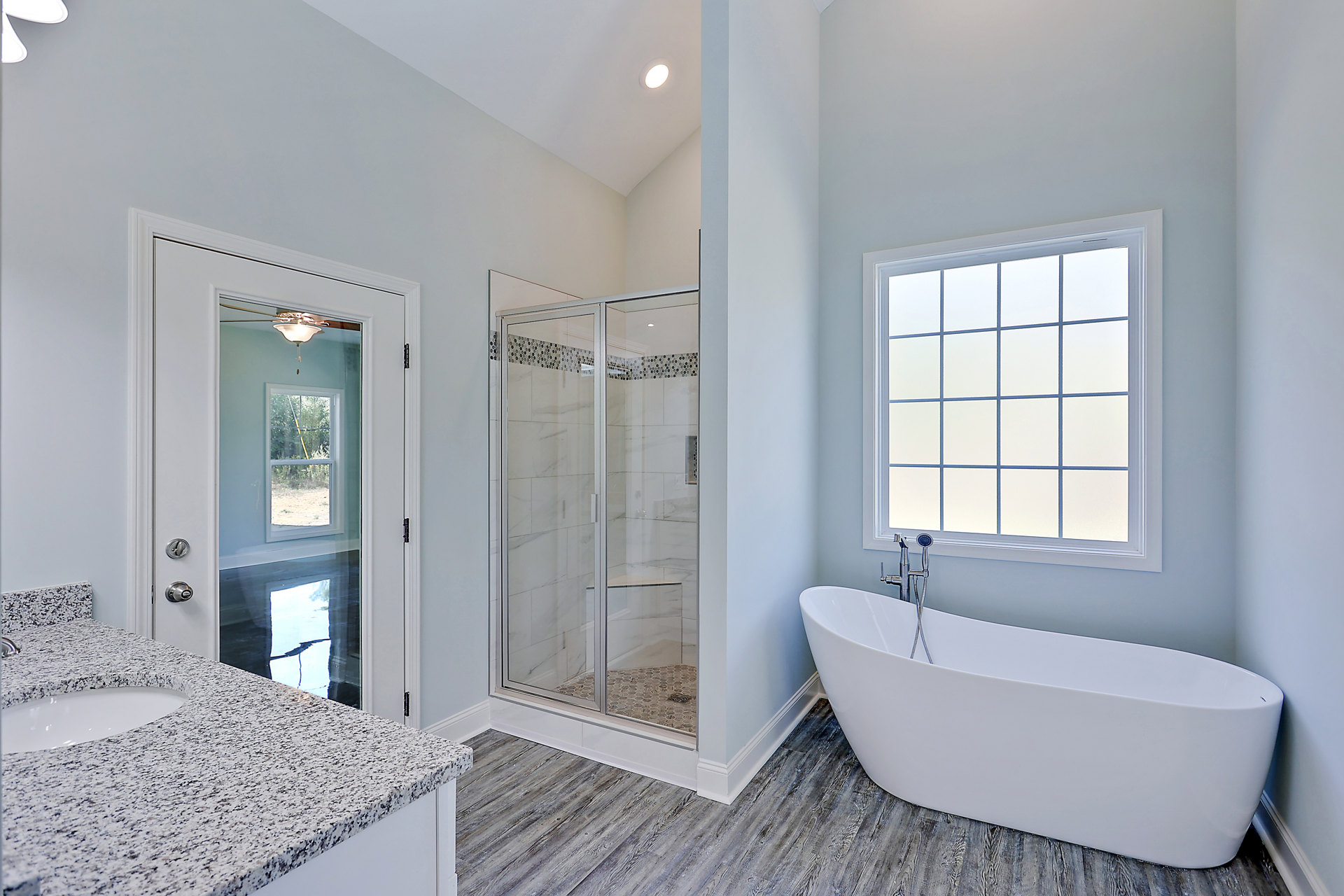 Modern bathroom featuring a white bathtub with silver fixtures, glass-enclosed shower, light-colored tile walls, countertop with sink, multi-pane window, and ceiling fan with