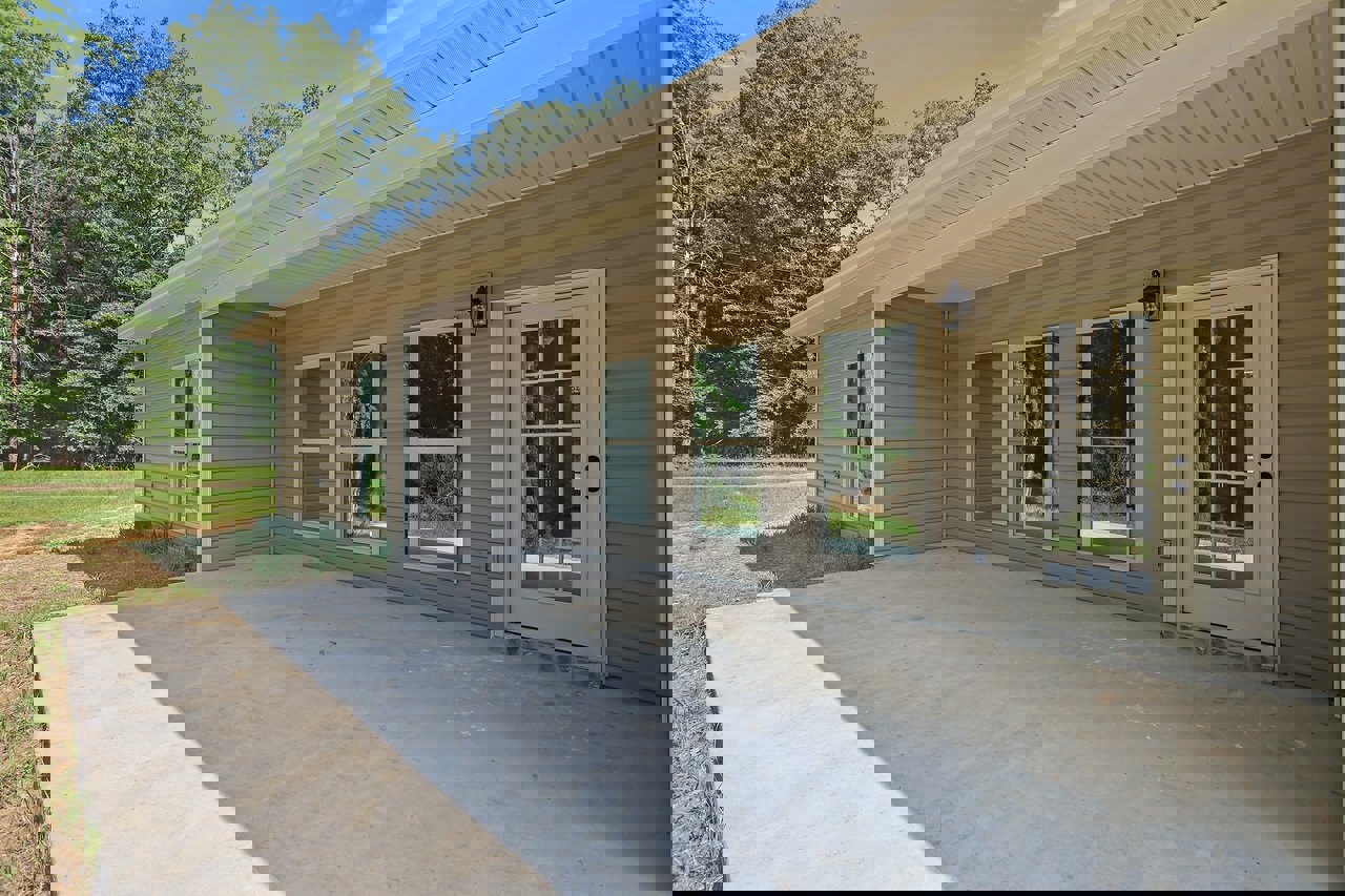 White siding house with covered porch, glass paneled front door, multiple windows reflecting trees, concrete patio, shaded by nearby trees.