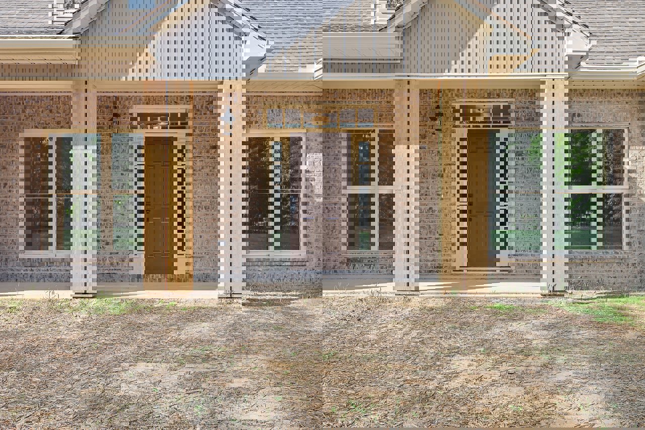 Brown front door with glass panes and metal handle, set in a brick exterior next to a white-framed window, with dirt landscaping in foreground.