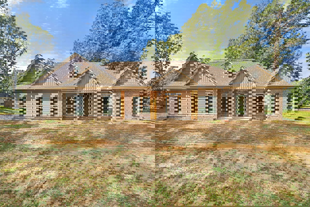 Two-story home with white siding, brown front door, white-trimmed windows, dirt patch in front yard, surrounded by grass and mature trees under a partly cloudy sky