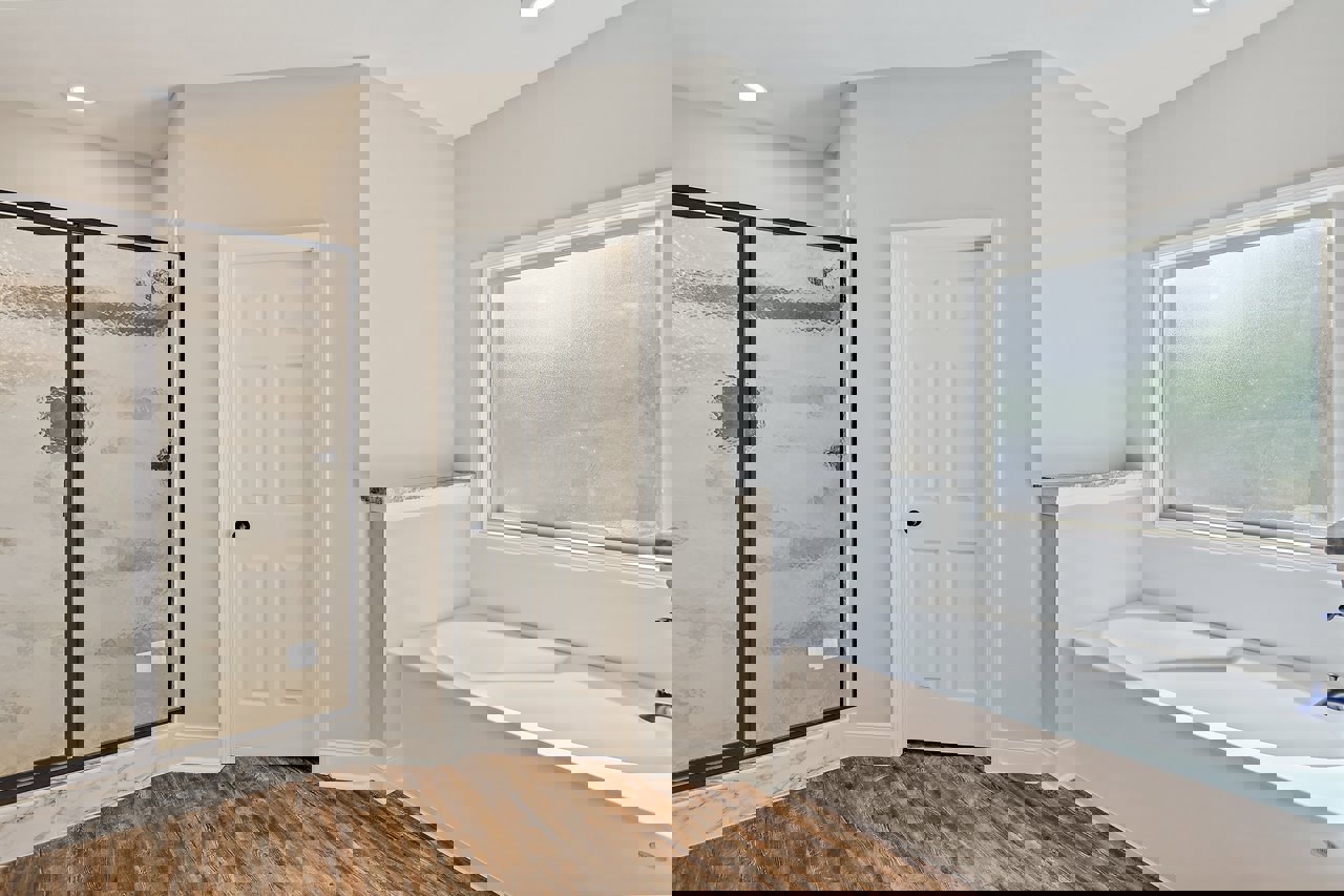 Bathroom with freestanding bathtub, glass-enclosed shower, frosted window, wood flooring, white walls, and white door with black handle