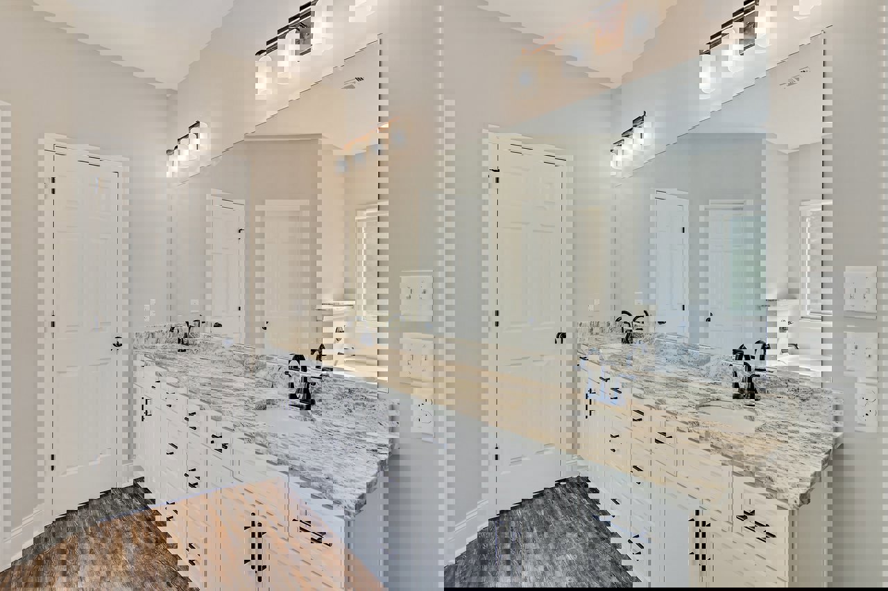 Bathroom with wide mirror above double sinks, white cabinetry with black handles, white walls, ceiling light fixture, and visible light switch.