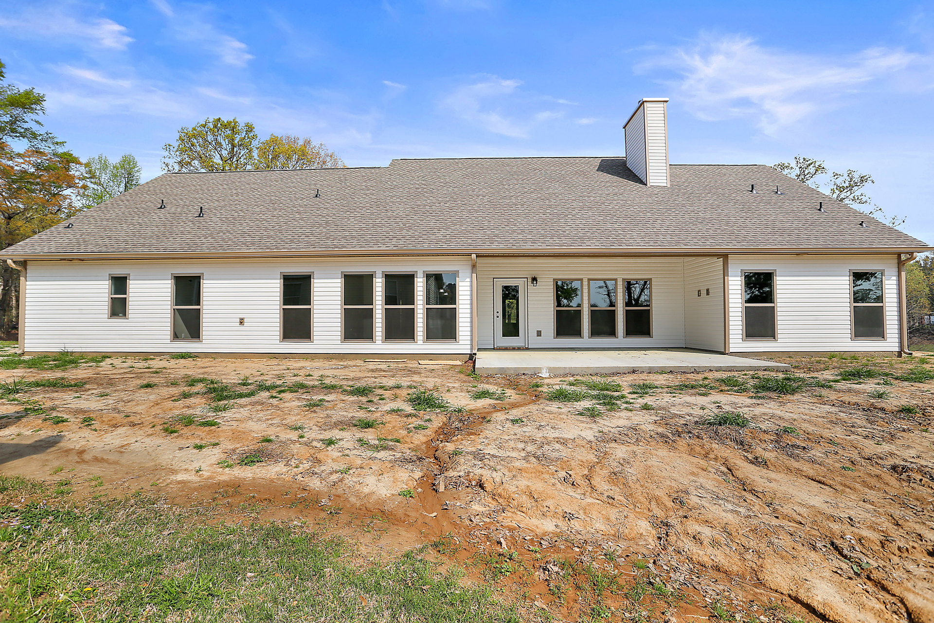 White house with rectangular windows and white door, dirt yard in foreground, Wyckoff House visible in background, trees reflected in window glass, cloudy sky overhead.