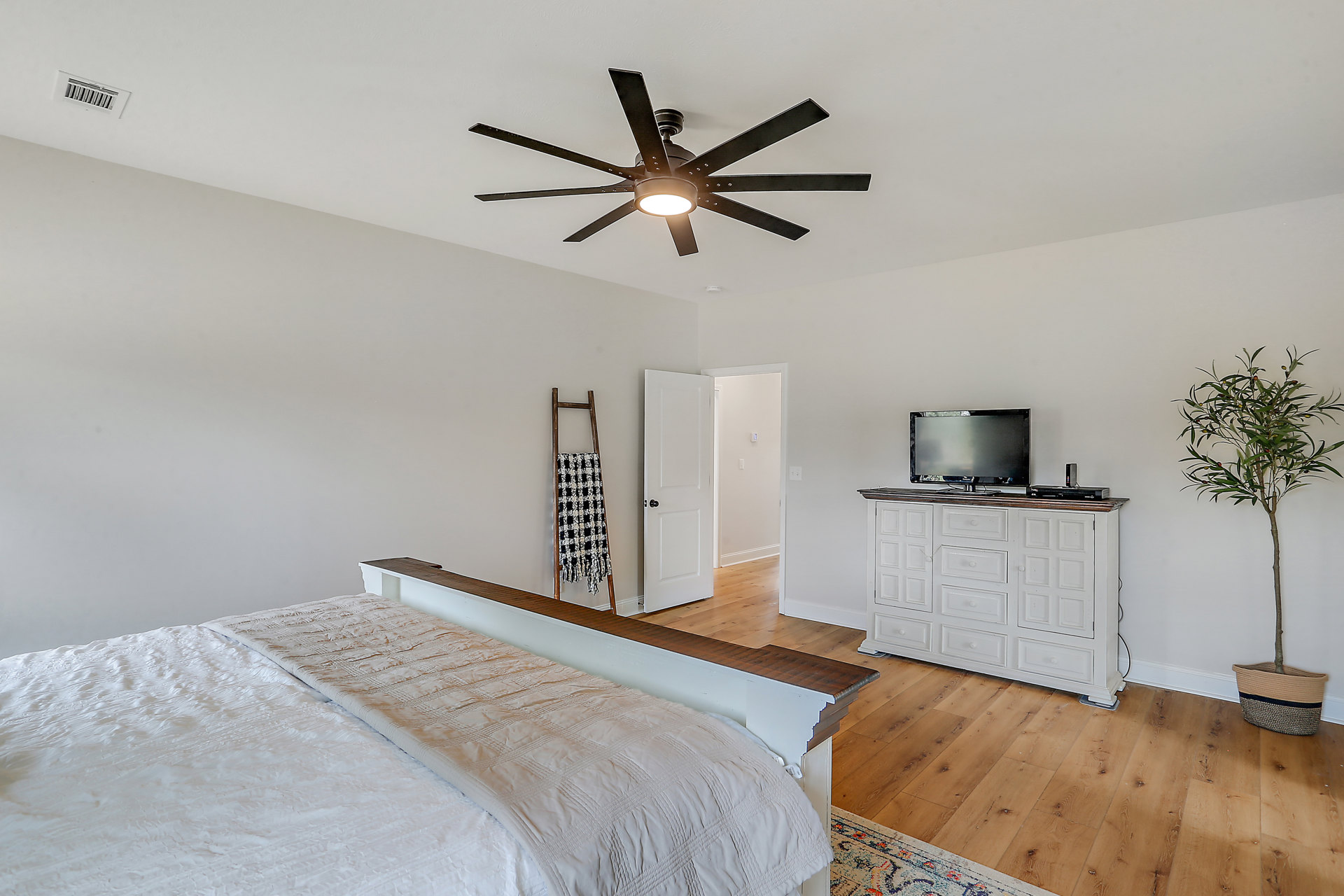 Bedroom with a ceiling fan, bed covered in a white blanket, black and white checkered fabric, wall-mounted TV, and leafy tree visible through window.