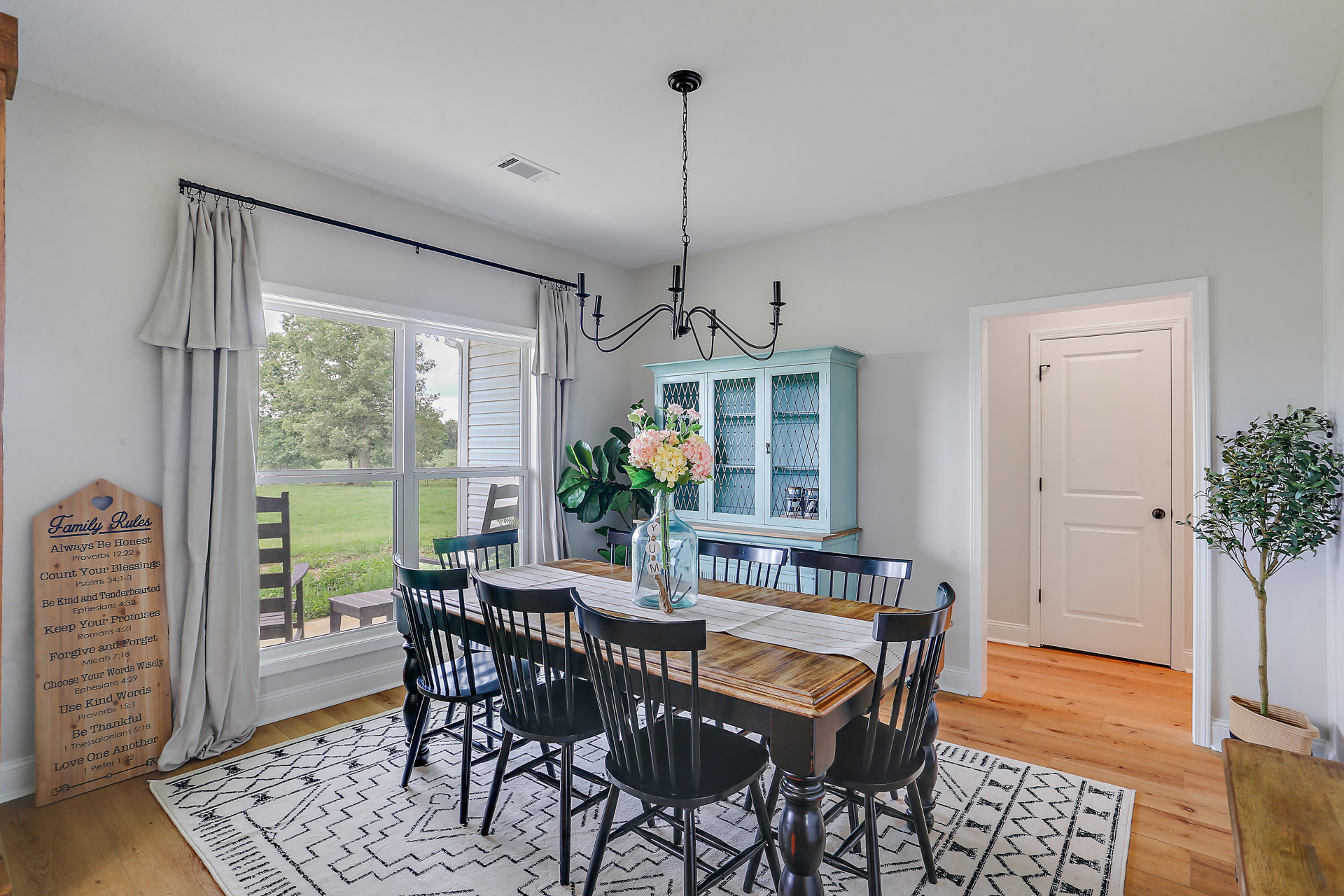 Dining room with wood table, black chairs, a vase centerpiece, potted tree, wood sign with black text, and white curtain along the wall