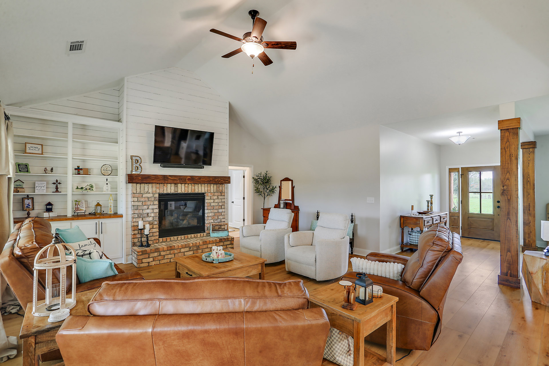 Living room with stone fireplace, brown leather sofa, white accent chair, ceiling fan with light, hardwood floors, and wall-mounted television