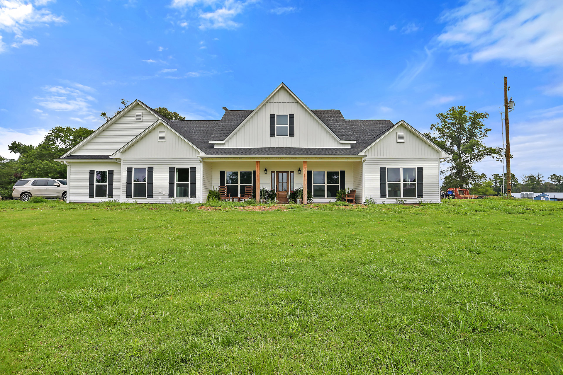 Two-story farmhouse with white siding, black shuttered windows, covered porch, large green lawn with scattered chairs, car parked on grass, Robert Frost Farm visible in the