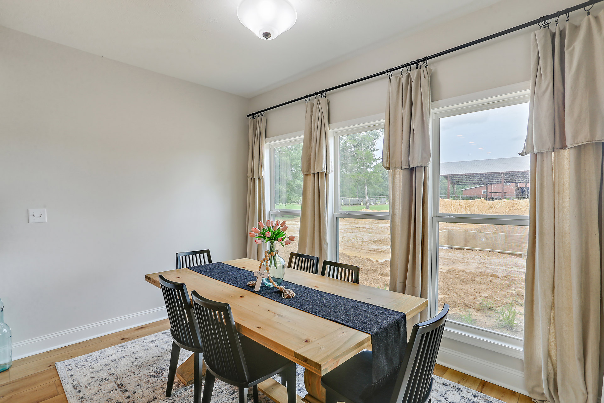 Wood dining table surrounded by chairs, topped with a vase of pink flowers and a wooden necklace, set near a window with sheer curtains and a visible wall light switch.