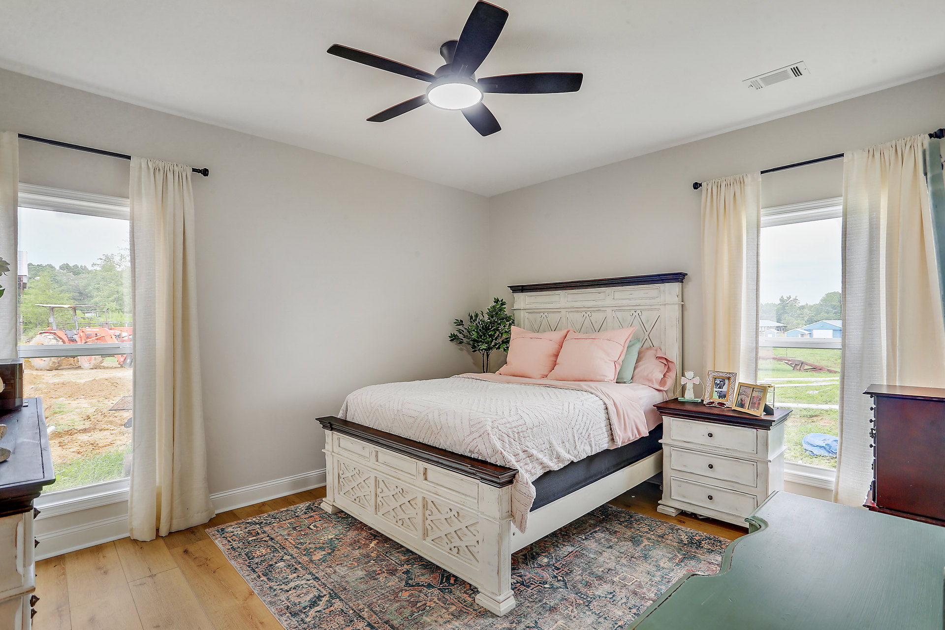 Bedroom with a ceiling fan, bed featuring pink pillows, white curtains, and a potted tree in the corner; red truck visible outside window.