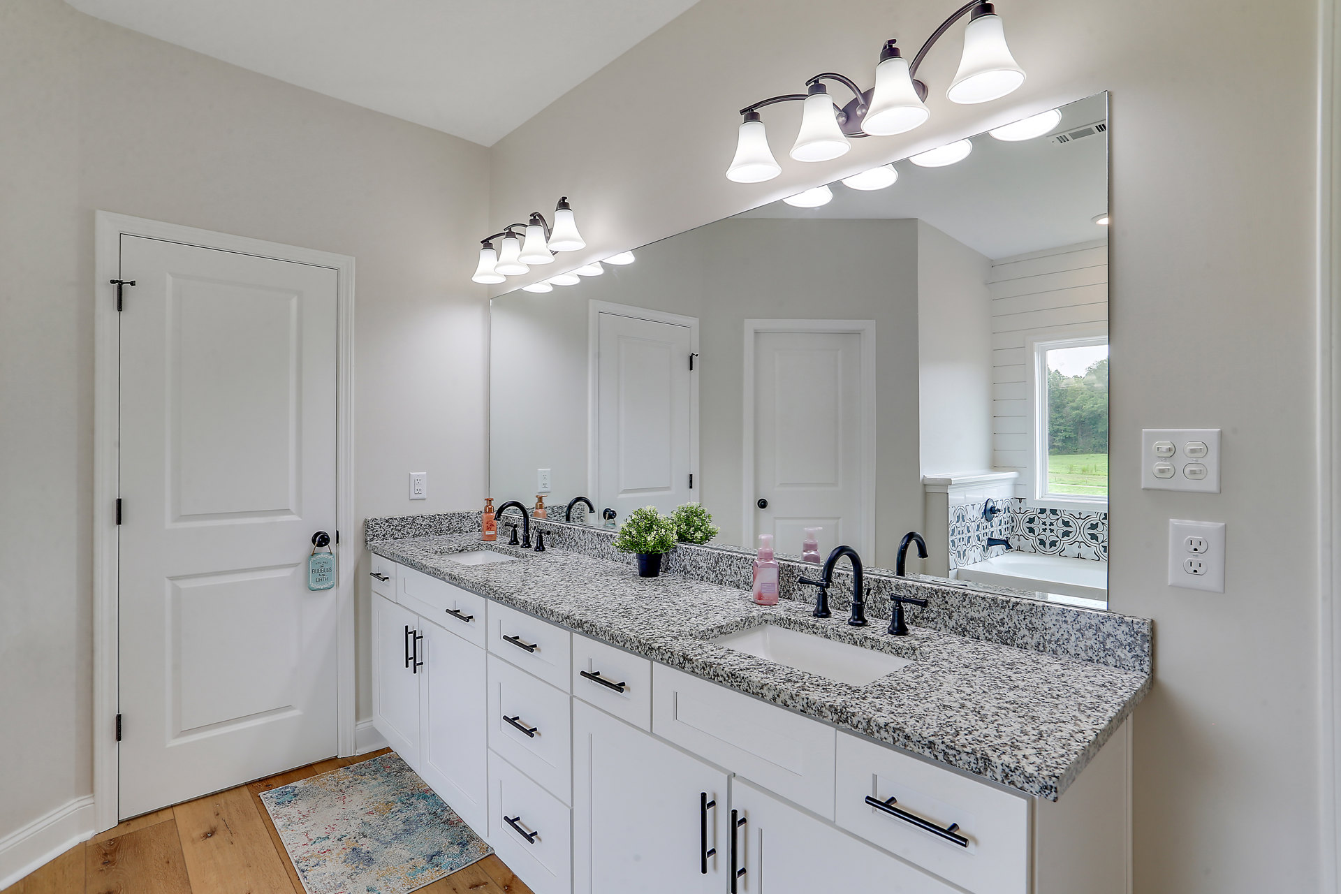 Bathroom with a wide framed mirror above a rectangular sink set in a stone countertop, white cabinetry below, wall-mounted light fixture with multiple bulbs, white door with a