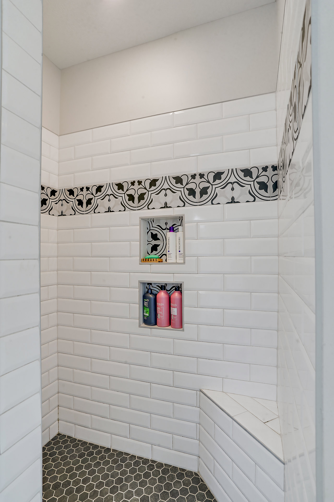 Shower featuring white tile walls with a black and white tile border, black and white tile floor, built-in shelf stocked with shampoo and soap bottles