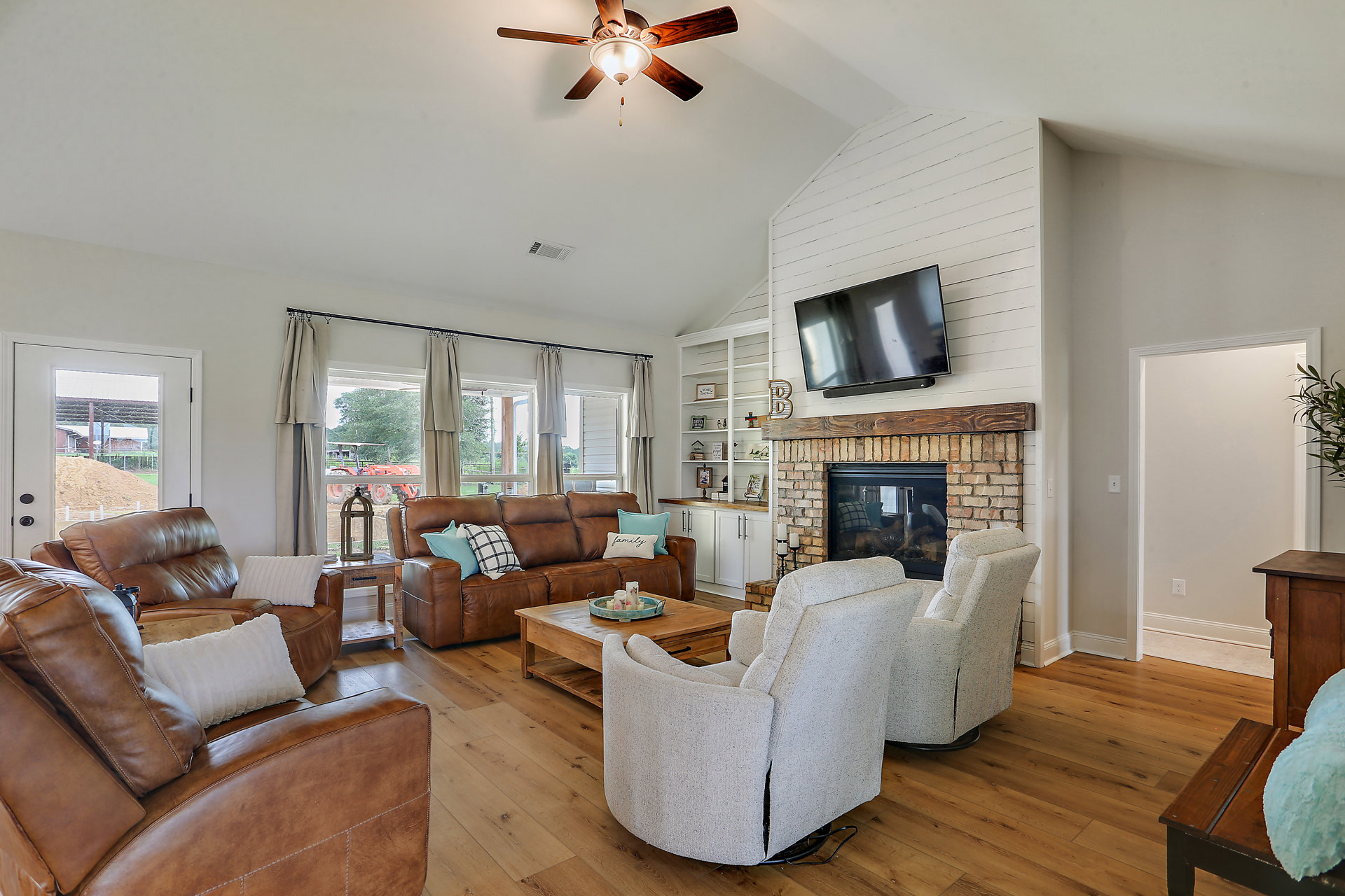 Living room with stone fireplace, mounted TV, gray couches with patterned pillows, white recliner, wood flooring, ceiling fan with light fixture, neutral walls