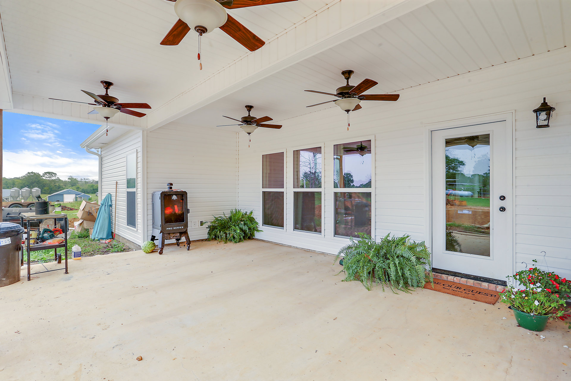 Covered porch featuring ceiling fans, stone fireplace, potted plants with red and white flowers, striped green foliage, blue curtain draped from a post, and glass window in a grey