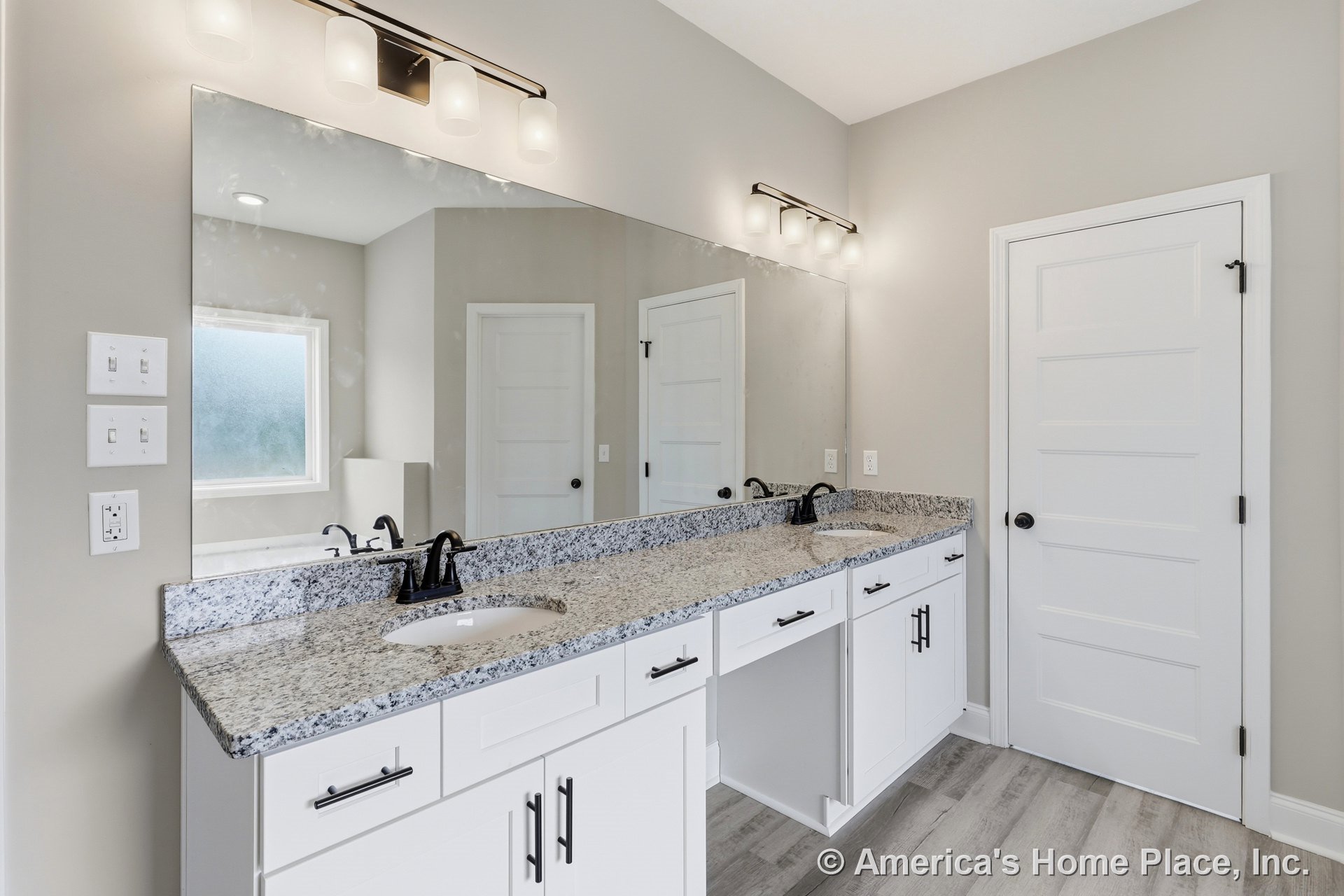 Spacious bathroom featuring a double sink vanity with white cabinetry, large frameless mirror, frosted window, white doors with black hardware, and light-colored tile flooring.