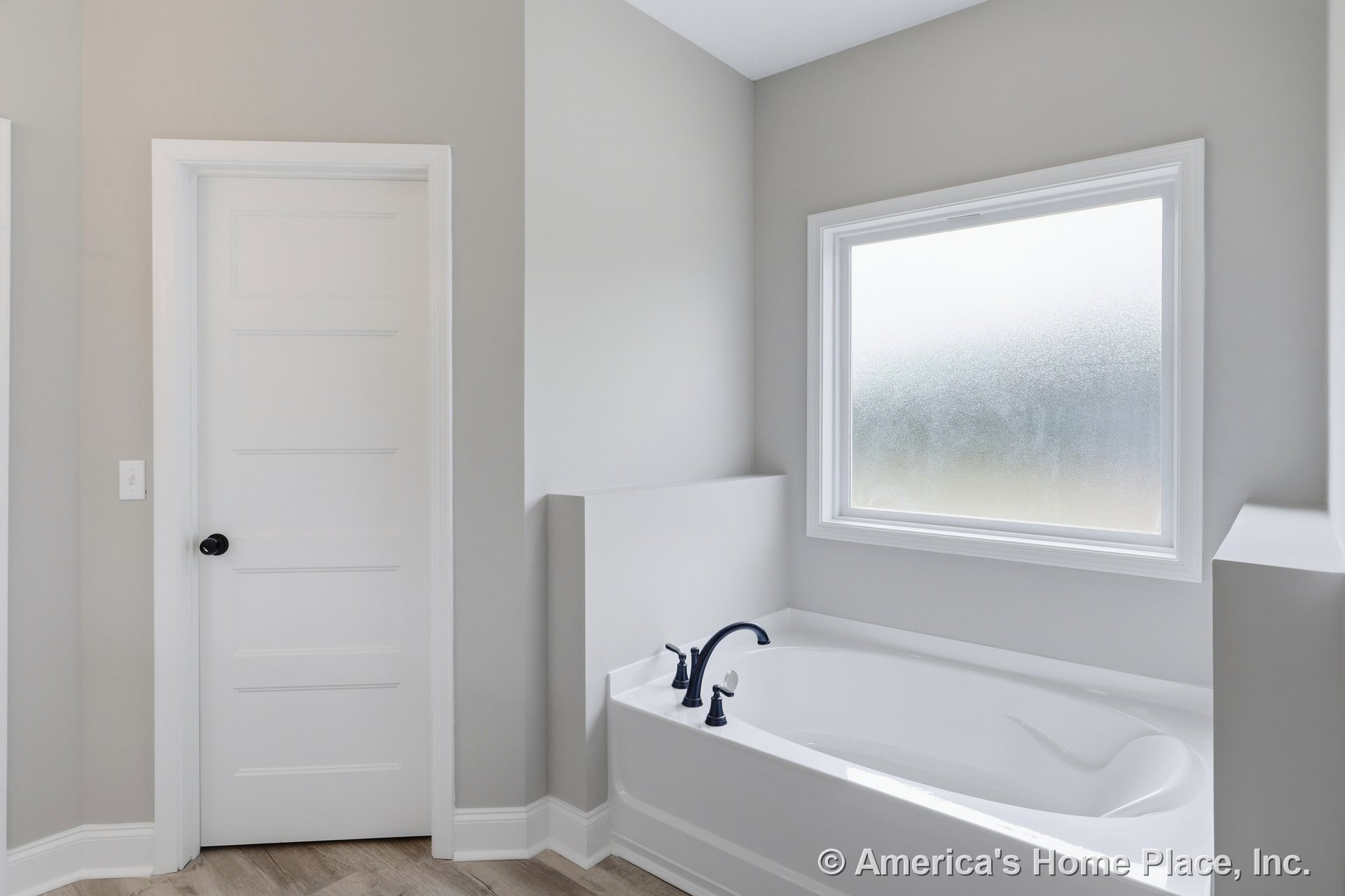 Freestanding white bathtub with chrome faucet beside frosted glass window, white walls, black trim, and white door with black knob