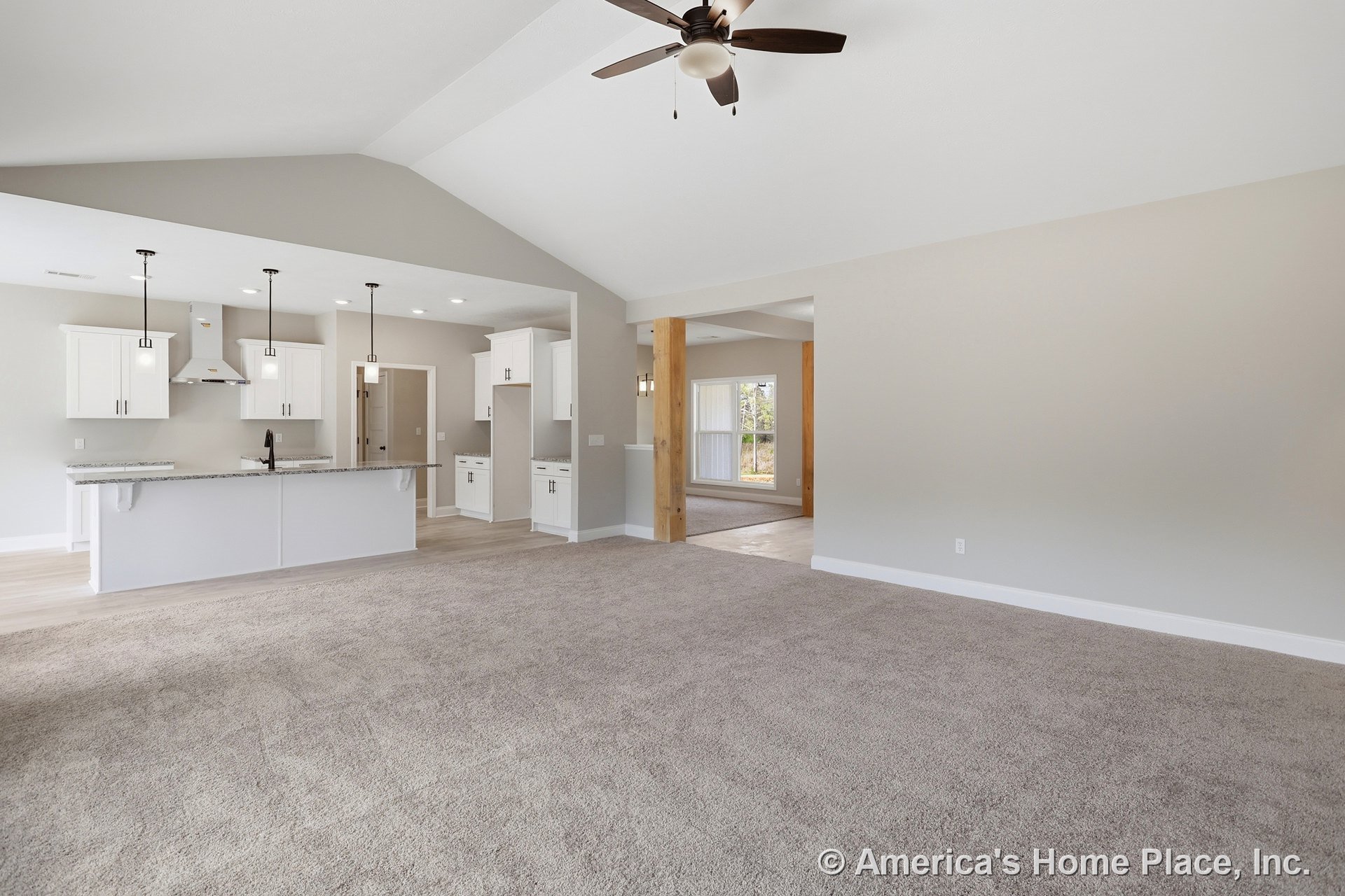 Open living room with beige carpet, ceiling fan with light, adjacent kitchen featuring white cabinets with black handles, kitchen counter with sink, and window with white frame