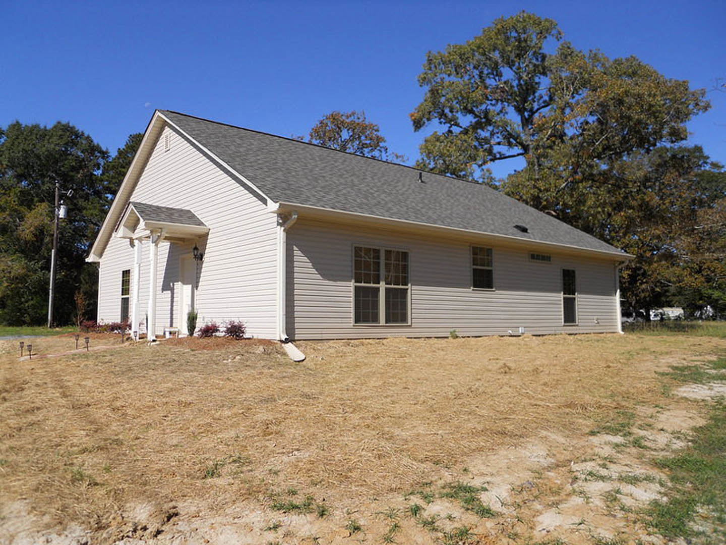 White siding house with square window, grassy front yard, pole-mounted light, mature trees in background