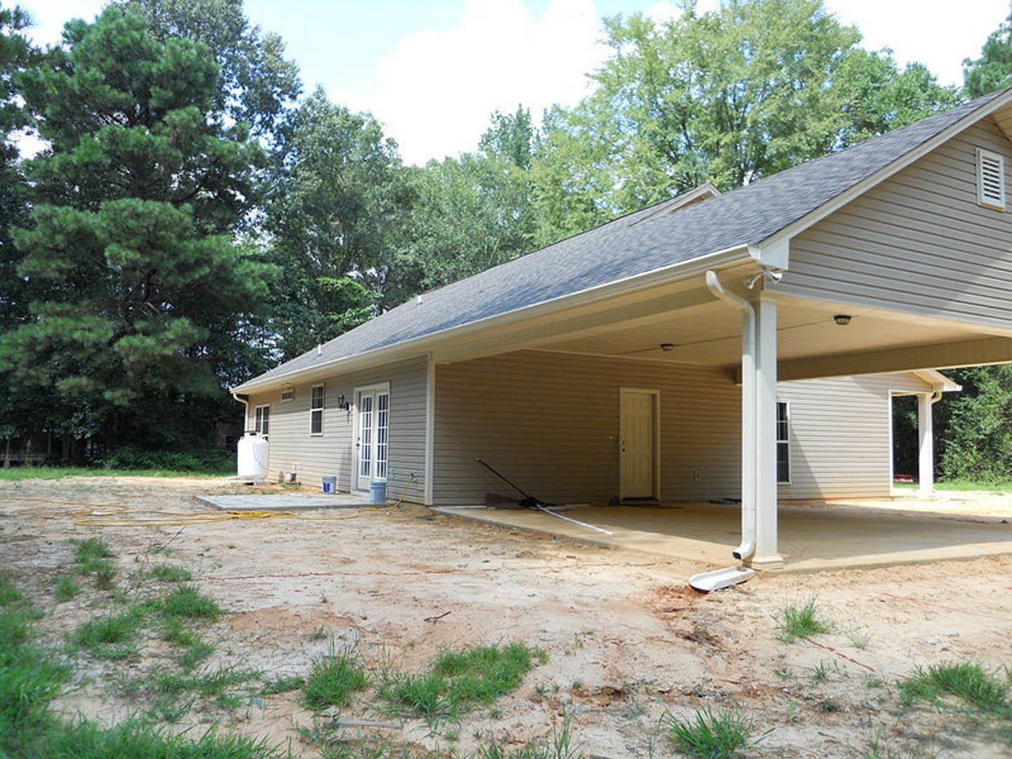 Covered front porch with white door, attached garage, dirt and grass yard, garden hose, leafy trees, blue sky
