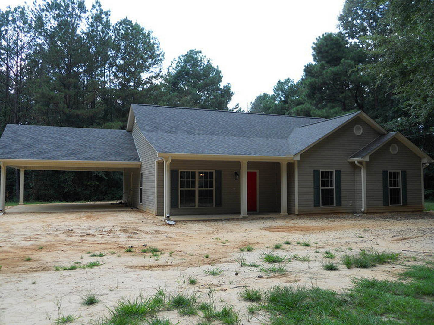 Two-story home with covered front porch, red entry door, white-framed windows, gray shingle roof, and patchy grass over dirt yard, surrounded by mature trees.