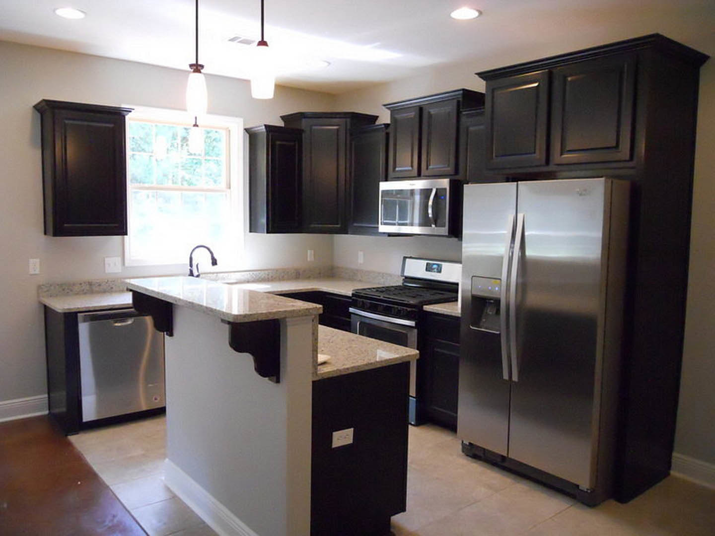 Black kitchen cabinets, stainless steel refrigerator and microwave, white countertop, window letting in natural light.