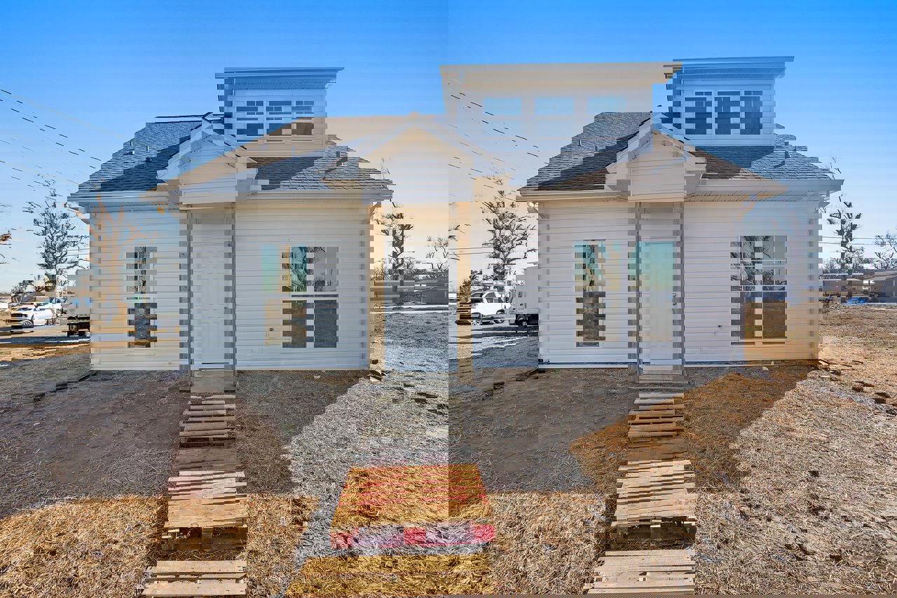 Two-story house with gray siding, covered front porch, white door with silver knobs, wooden pallet on the lawn, wooden fence with stacked logs, white car parked on street, leafy