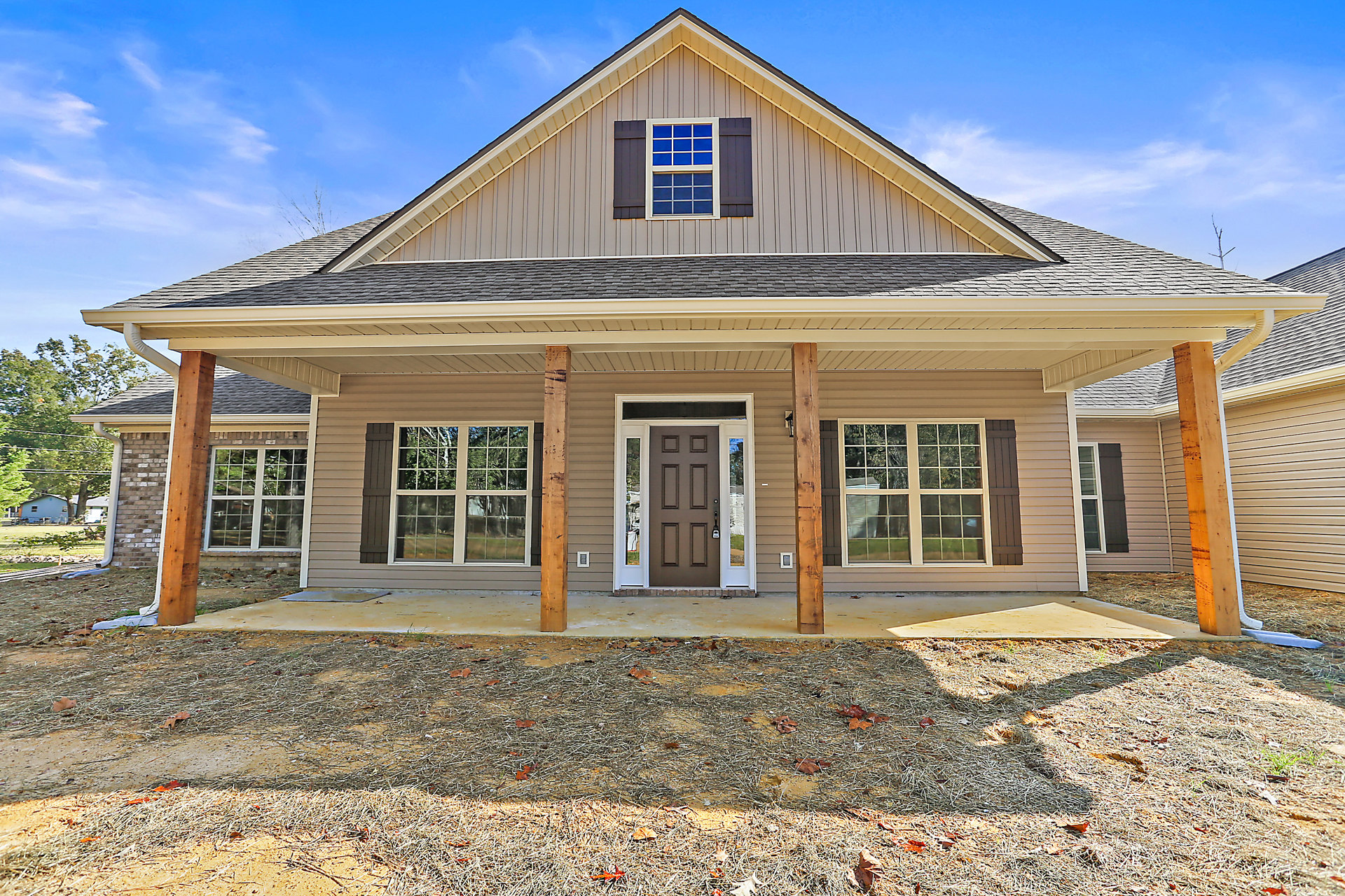 Large front porch with white columns, brown door framed in white, multiple windows reflecting trees and sky, light-colored siding, landscaped front yard