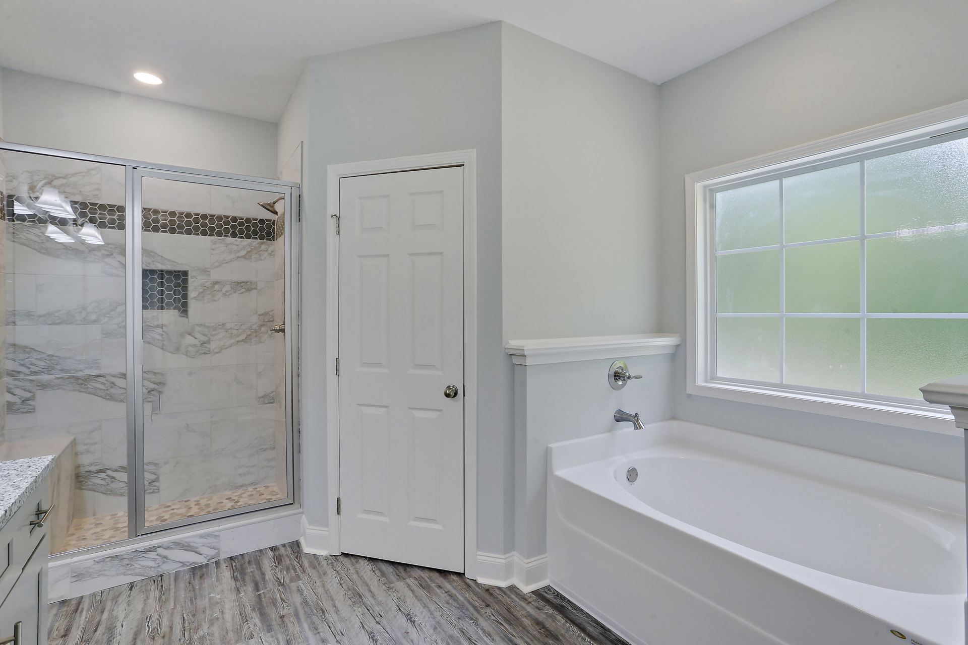Modern bathroom featuring a freestanding bathtub with chrome faucet, glass-enclosed shower, white paneled door with silver handle, frosted window, and light gray tile walls.