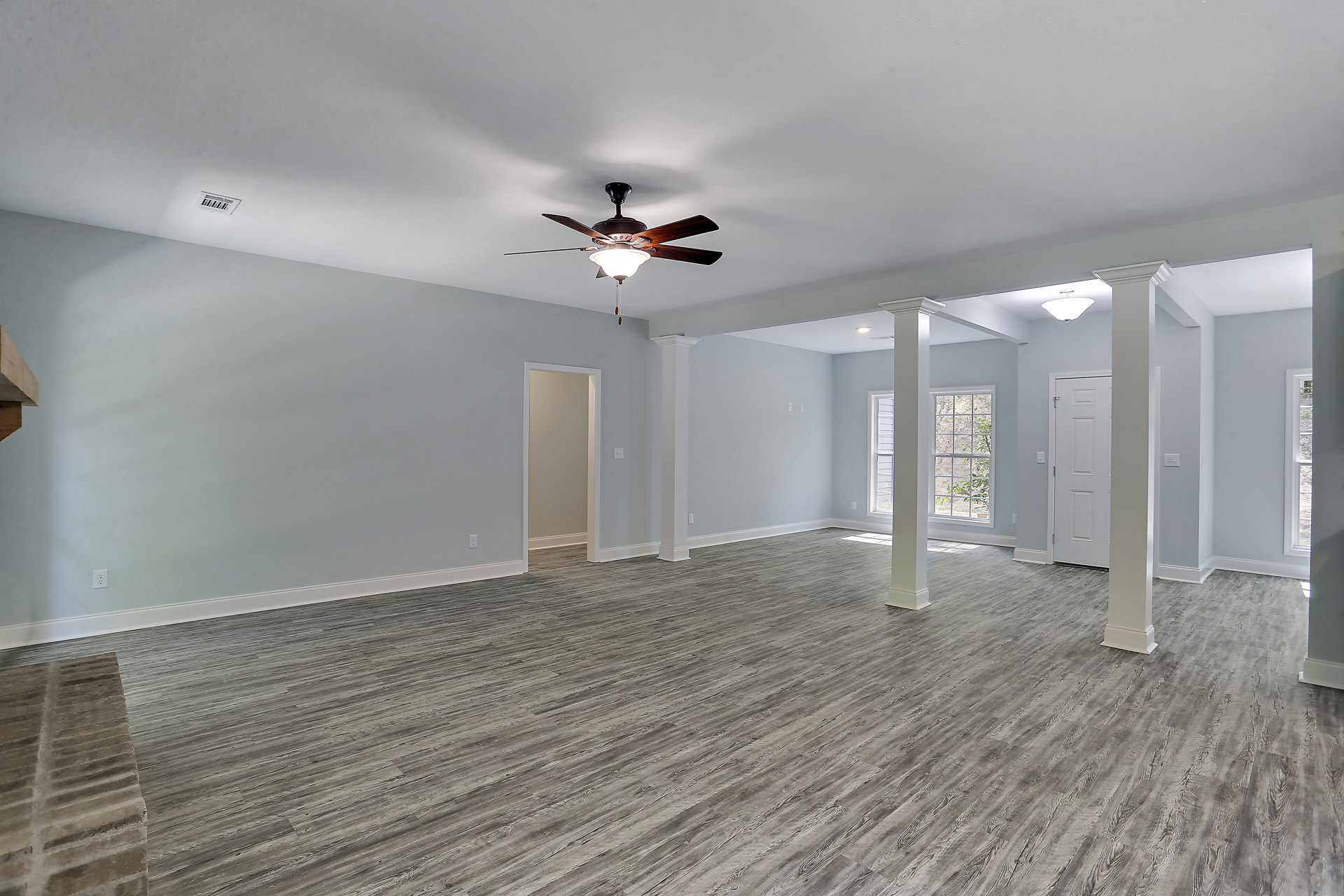 Open living space with wood flooring, white columns, ceiling fan with light fixture, white door, and window overlooking a tree.