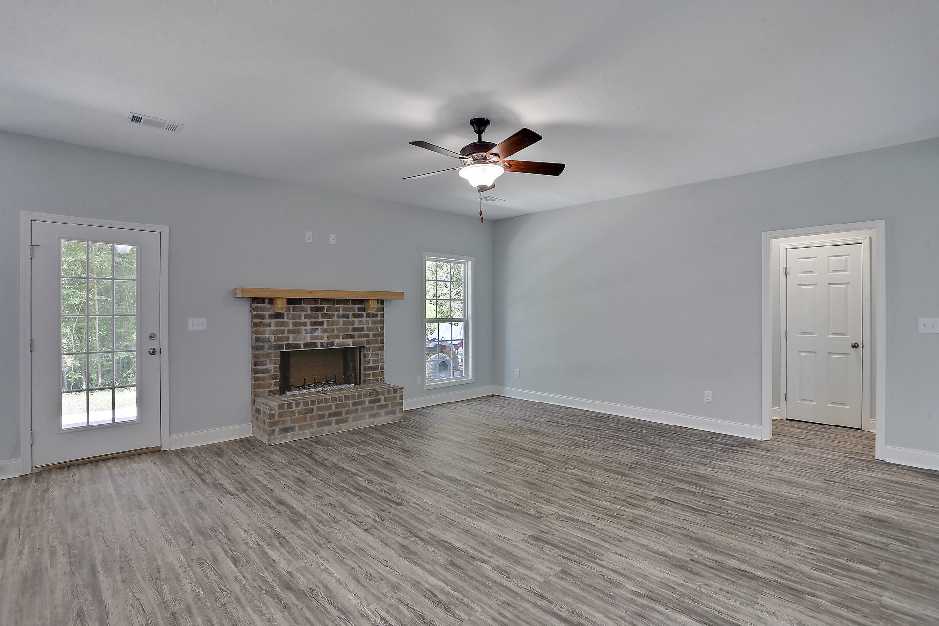Living room with wood flooring, plaster walls, ceiling fan with light fixture, fireplace topped by wood beam, glass-paneled door, and window overlooking trees and a tent.