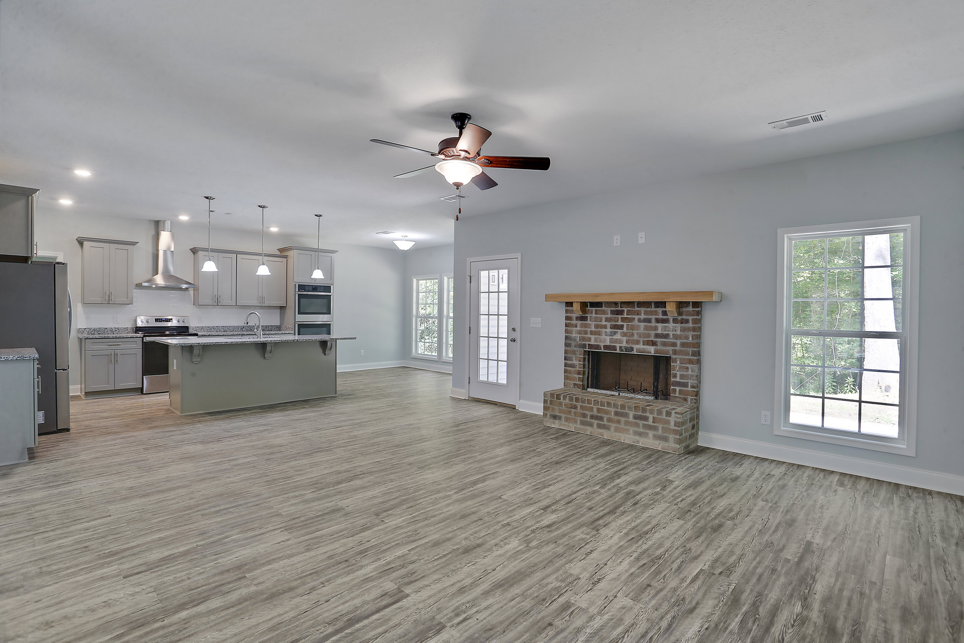 Open living room with brick fireplace, ceiling fan with light, adjacent kitchen, white door with glass panes, window overlooking trees, plaster walls, hardwood flooring