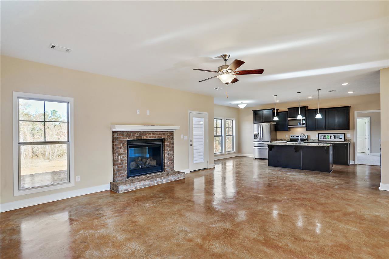 Living room with brick hearth fireplace, glass door, ceiling fan, brown wood flooring, large window showing trees, partial view of stainless refrigerator and cabinetry