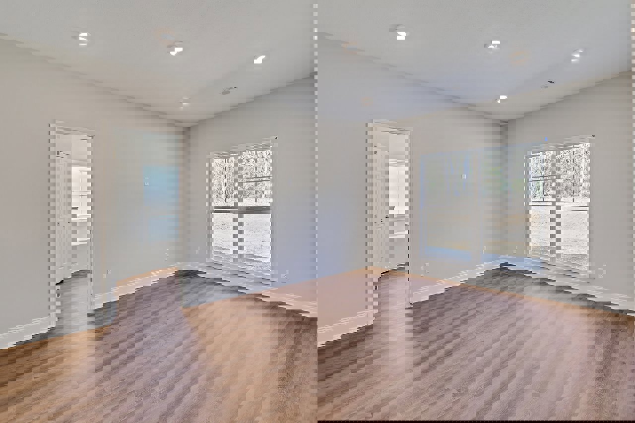 Wood flooring in a bright room with a large window overlooking trees, white walls, recessed ceiling lights, and a white door with silver knobs.