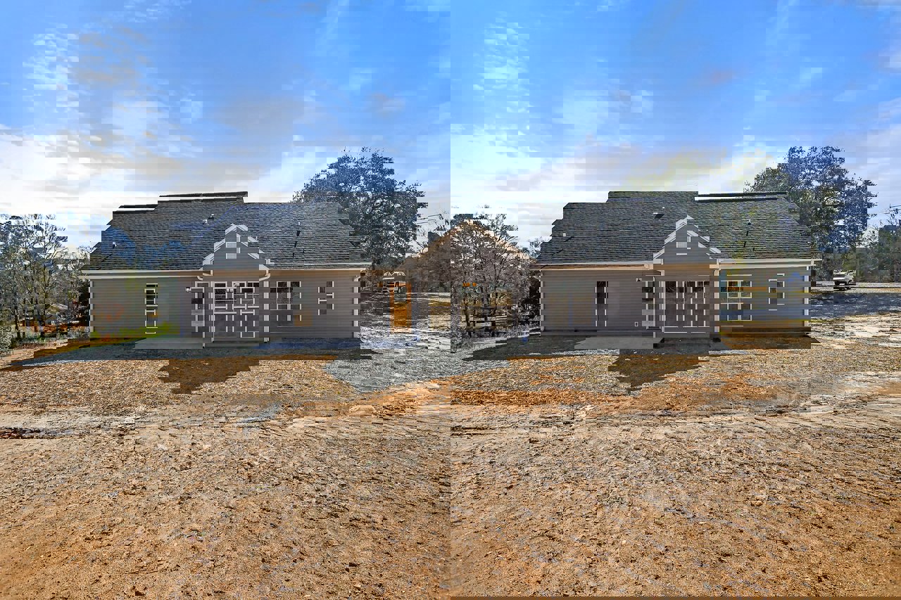 Partially built house with shingled roof, exposed framing, dirt lot, and surrounding trees under blue sky