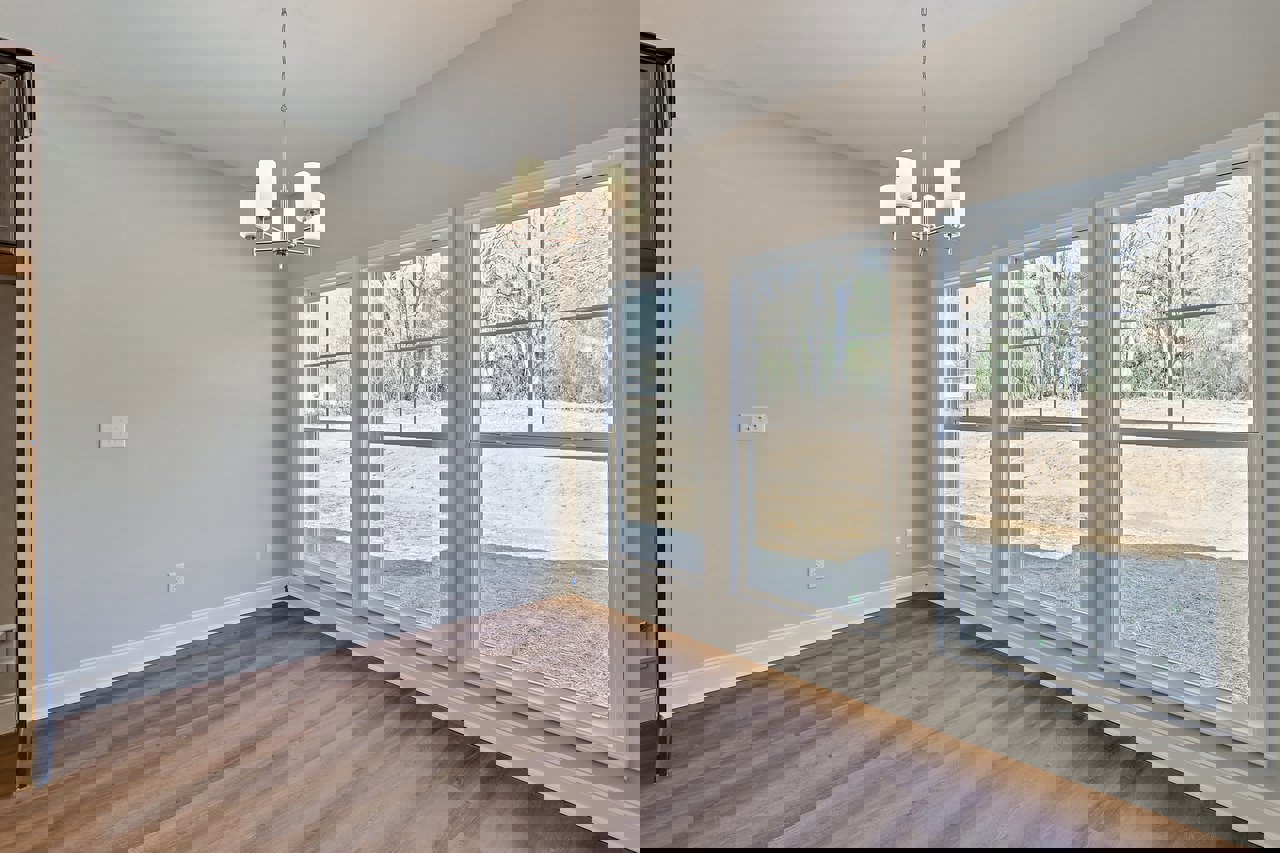 Chandelier with white shades hanging in a room with wood flooring, white walls, and large windows overlooking a dirt road