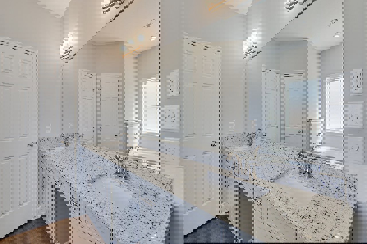 Bathroom with double white sinks and silver faucets, expansive wall mirror above, white cabinetry and drawers, tiled floor, window with white frame, white door with silver handle.