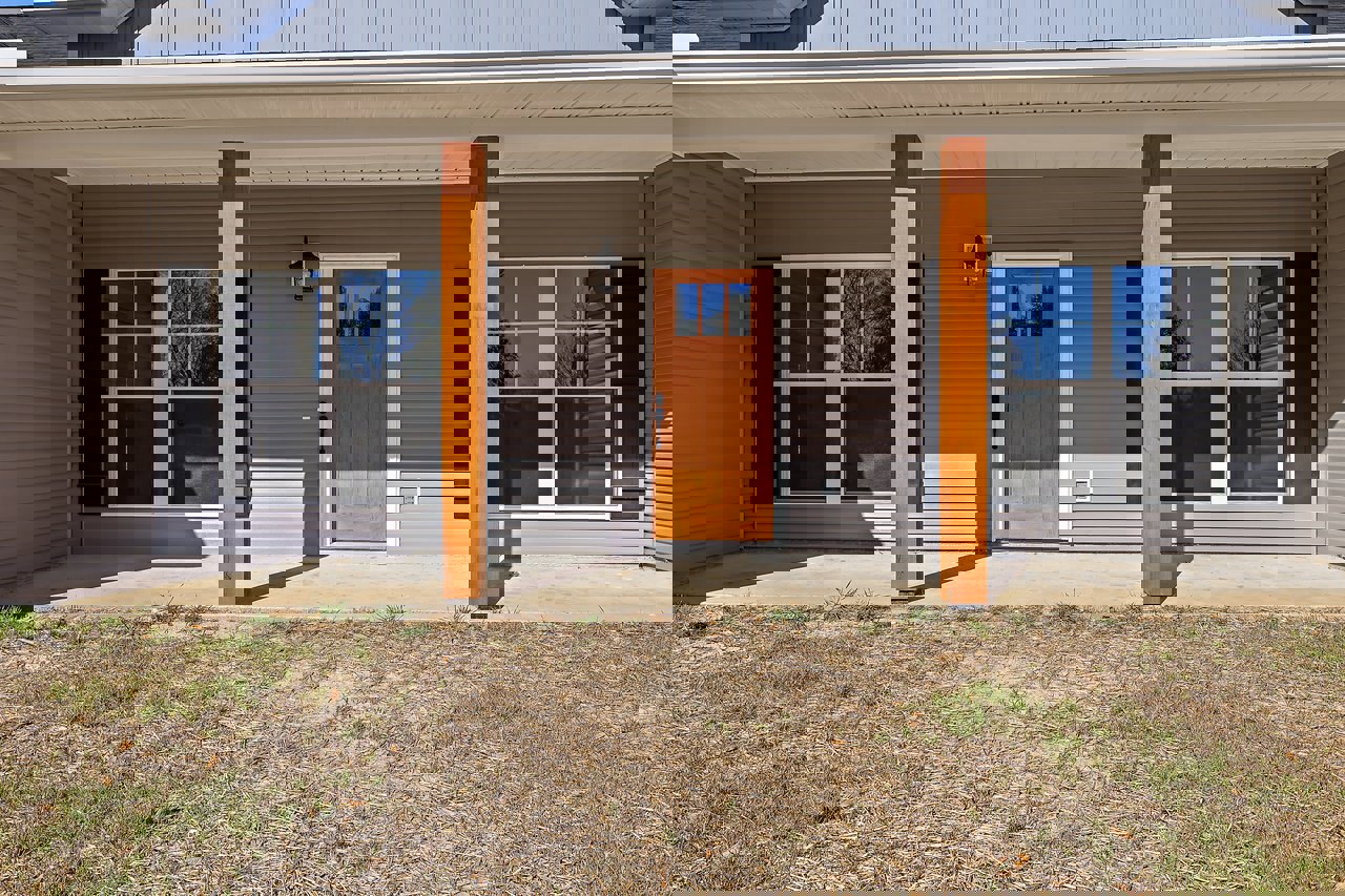 Front porch with wooden door featuring glass panel, wood posts, white siding, windows, and grassy yard with patches of dirt