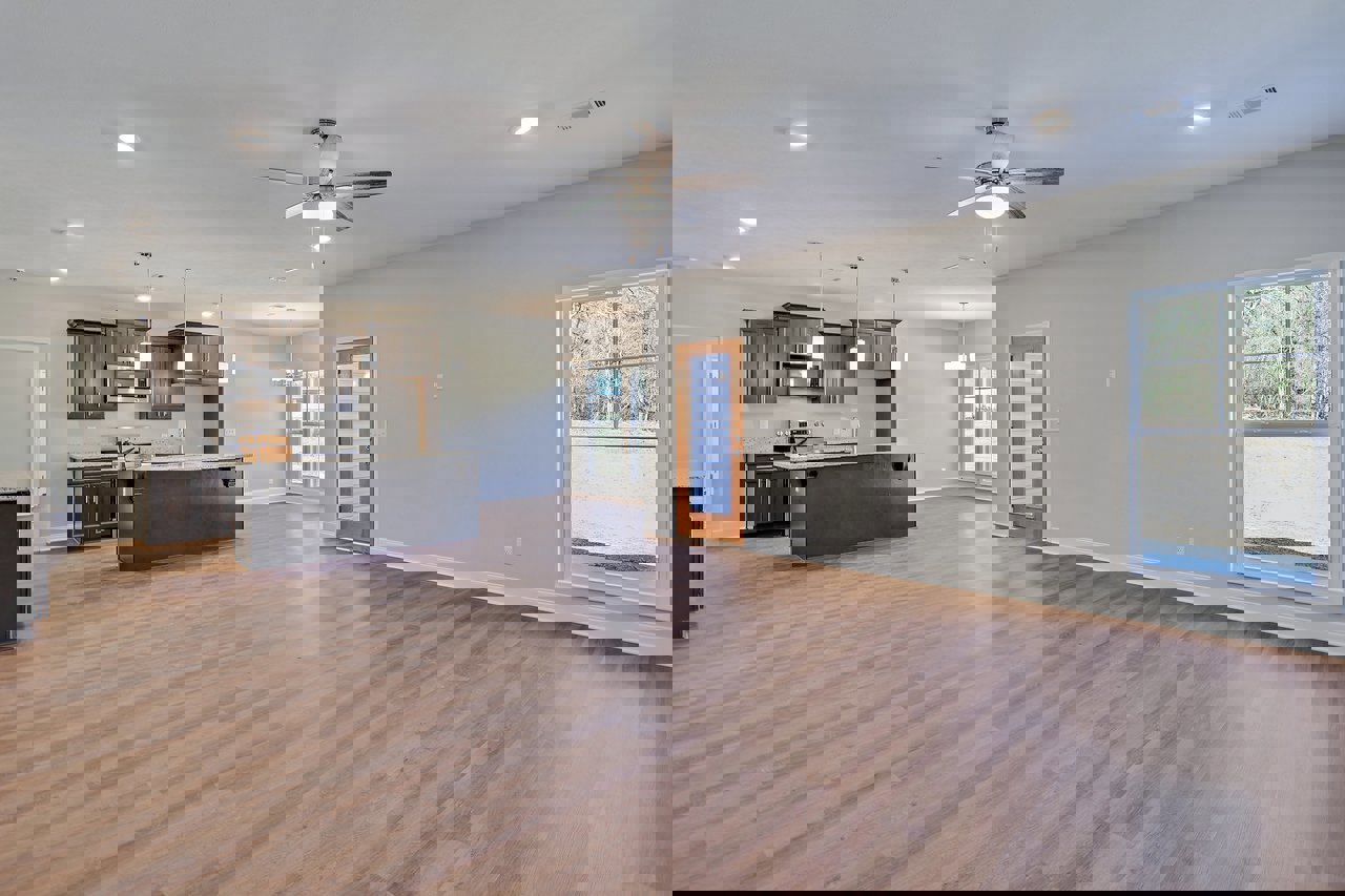 Spacious open floor plan featuring hardwood flooring, modern kitchen cabinetry, dining area, ceiling fan with light, large window overlooking trees, black accent wall, and a brown