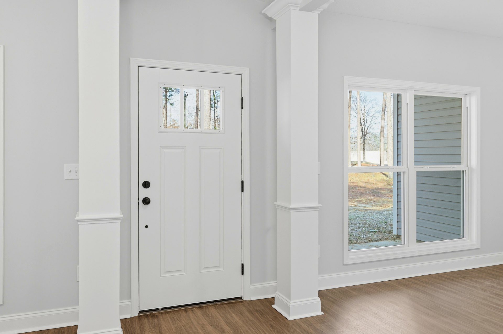 White paneled door with glass window, adjacent to a row of windows overlooking trees, white column, and light wood flooring