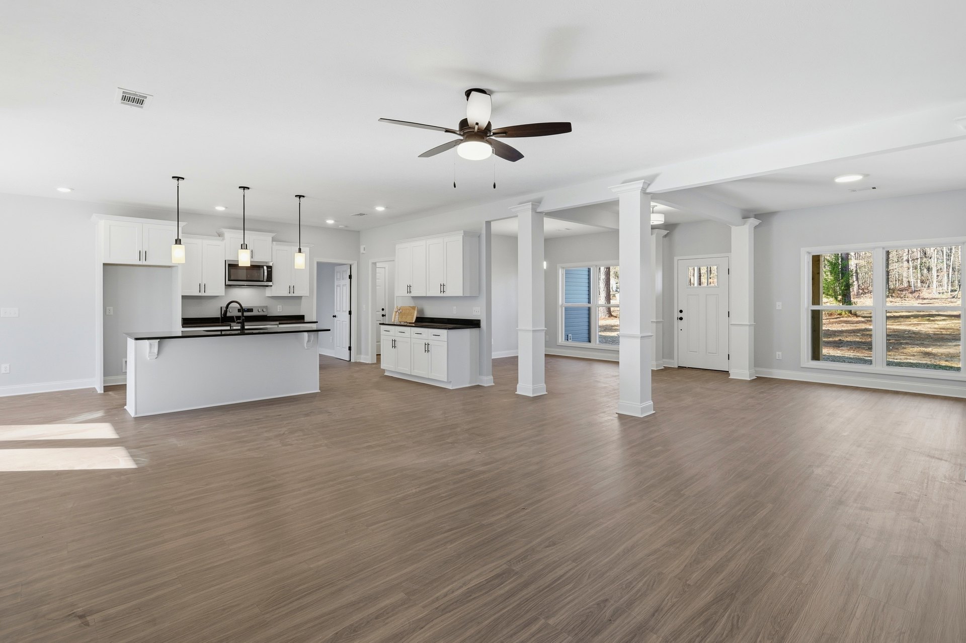 Spacious white living area with laminate wood flooring, ceiling fan with light, white columns, open kitchen featuring black countertops, large windows overlooking forest scenery