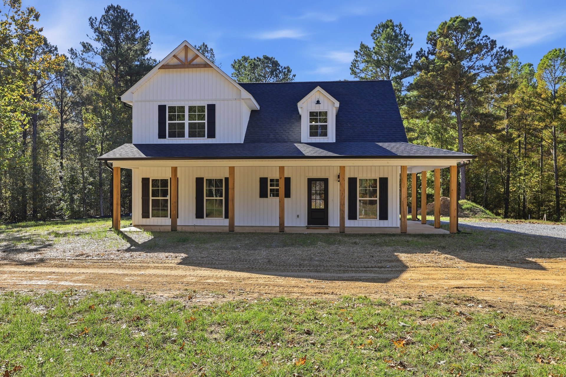Two-story house with blue metal roof, covered porch, white-framed windows, glass-paneled front door, dirt road leading to entrance, surrounded by mature trees and greenery.