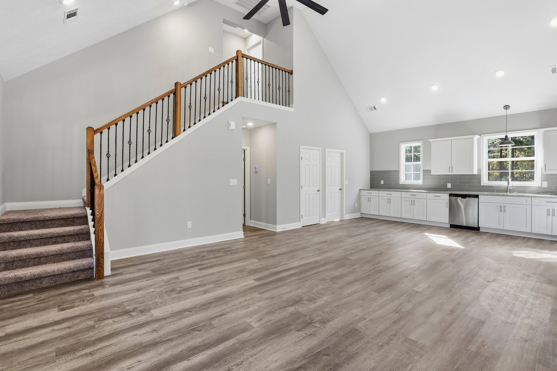 Open-concept living area with wood flooring, white walls, carpeted staircase with white railing, ceiling fan, and window with white trim