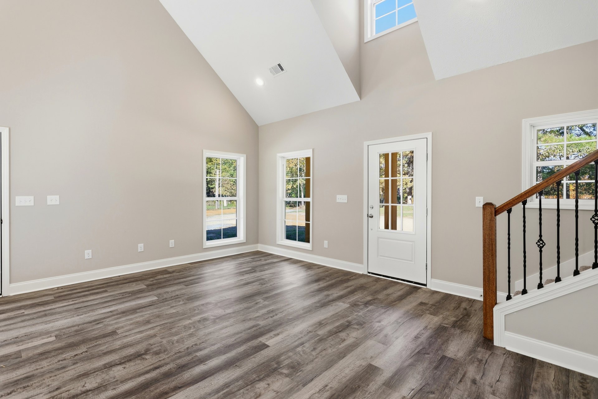 Wood floor room featuring a white door with mirrored glass, wooden staircase railing, plaster walls, and a window overlooking trees.