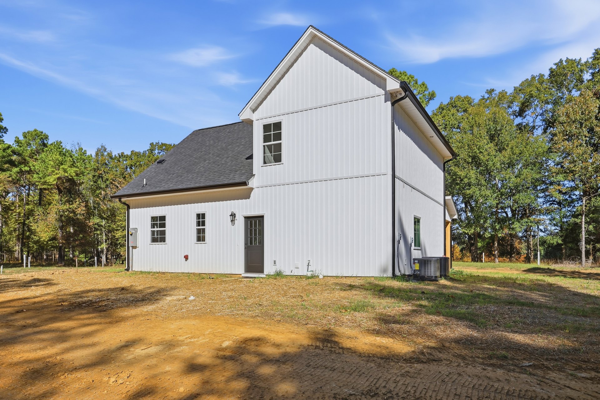 White siding house with black shingle roof, grassy yard, and dirt patch in front, surrounded by trees under a cloudy sky