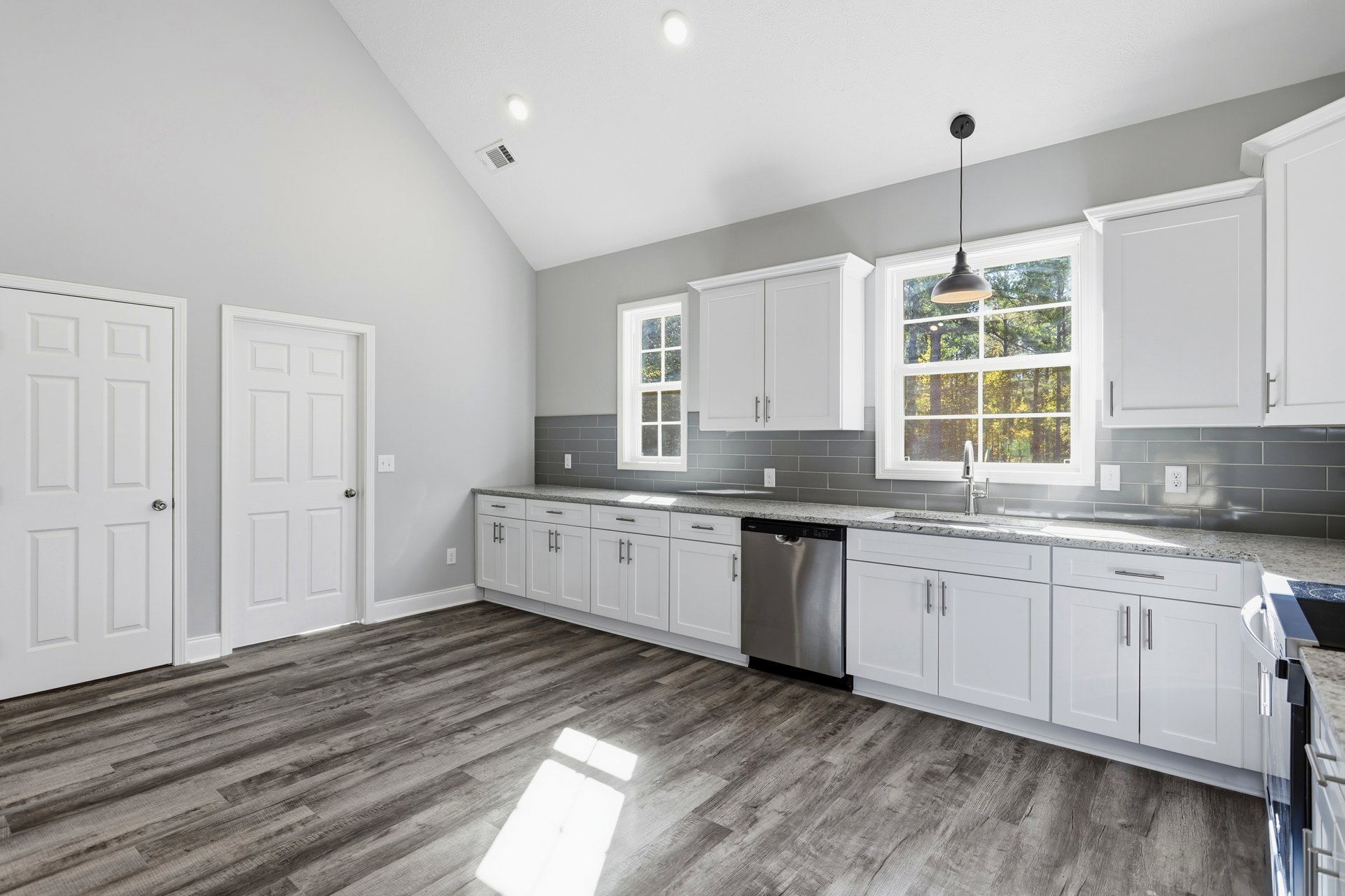 White kitchen cabinets with silver hardware, wood flooring, stainless steel dishwasher, white door with silver handle, ceiling light fixture, window above countertop
