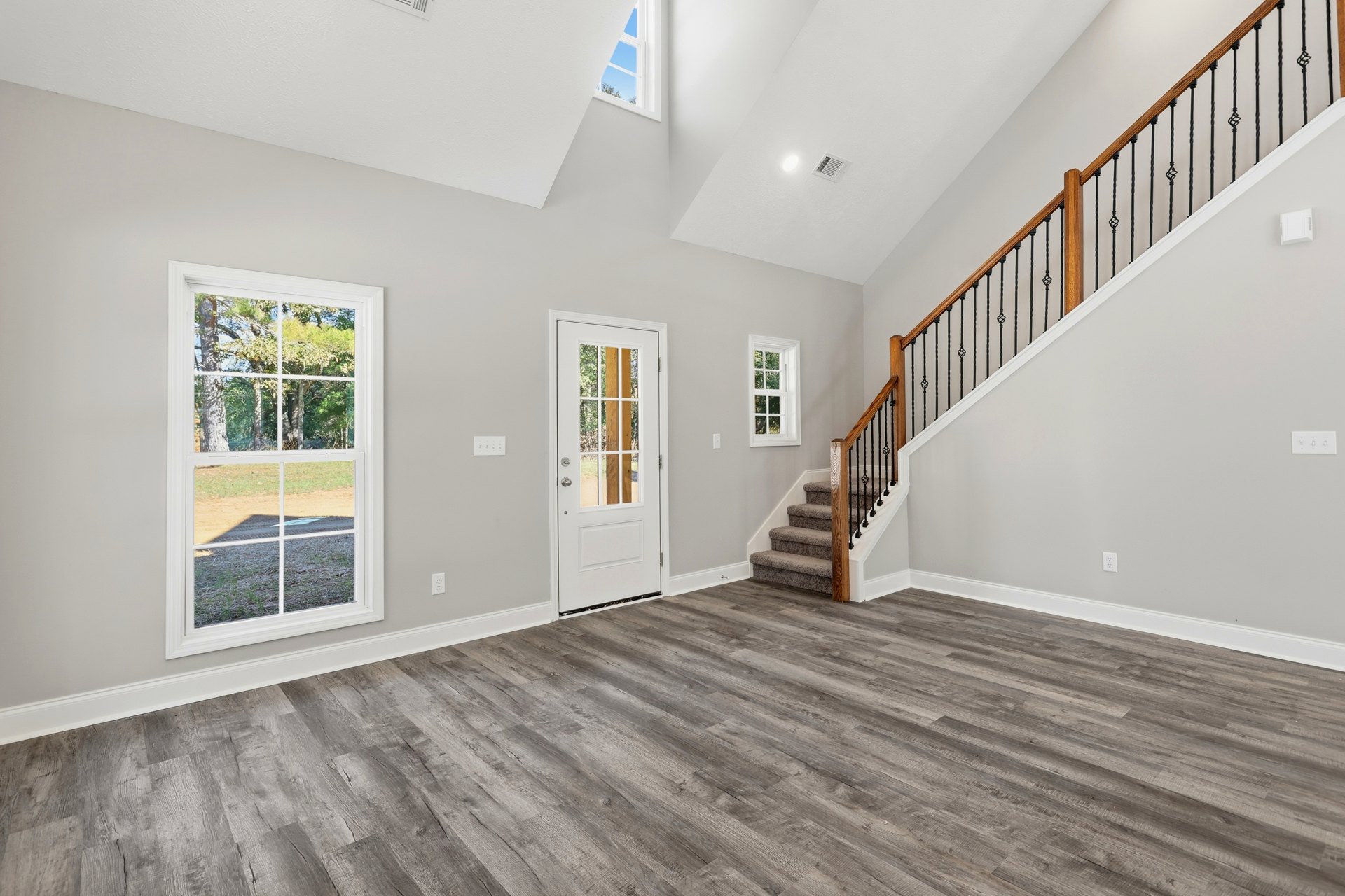 Hardwood floor room featuring a wooden staircase, large windows with white frames overlooking trees, and a white door with glass panes.
