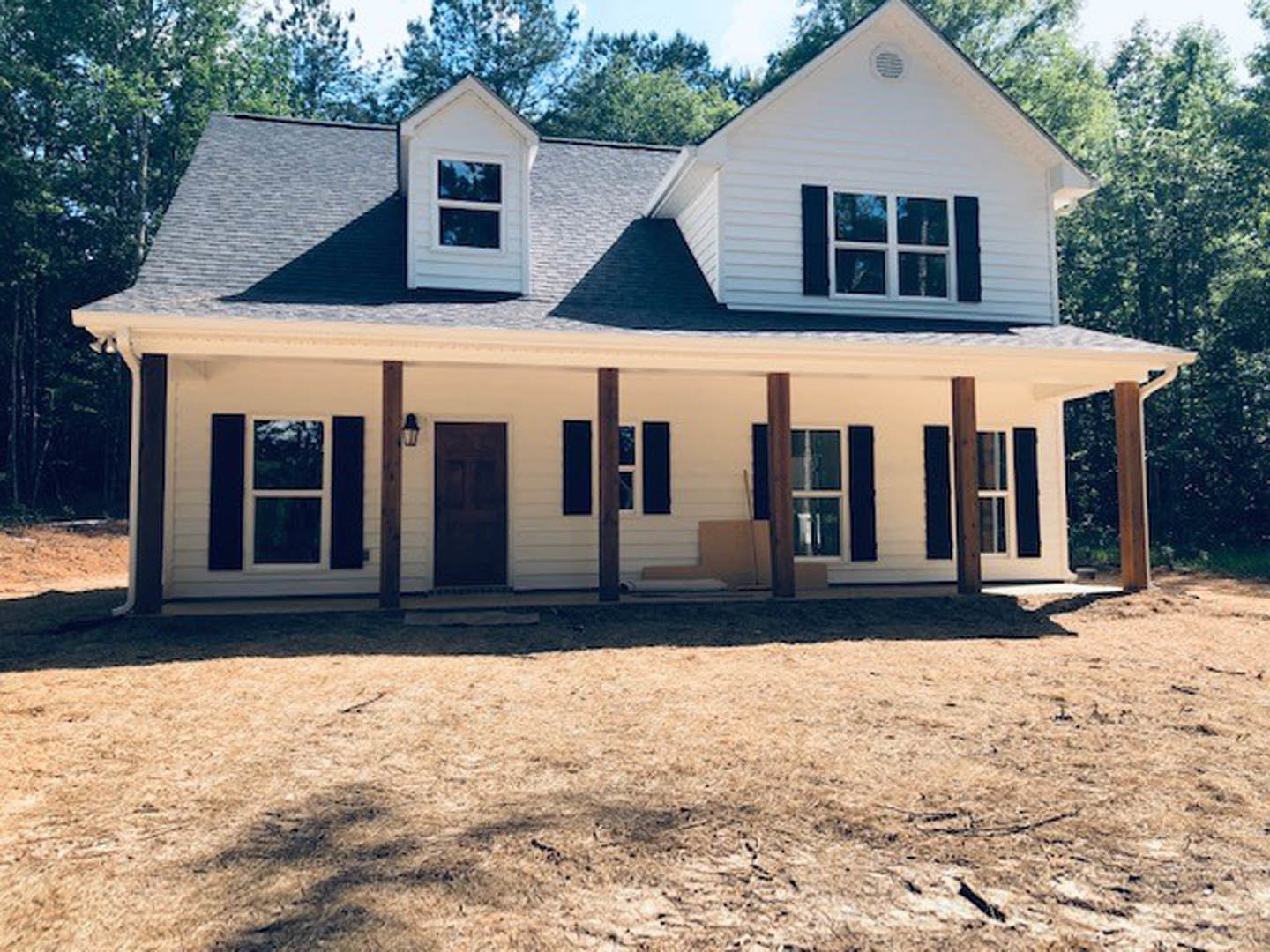 White house with wooden porch posts, black front door with white trim, large windows, dirt yard in foreground, tree and blue sky in background