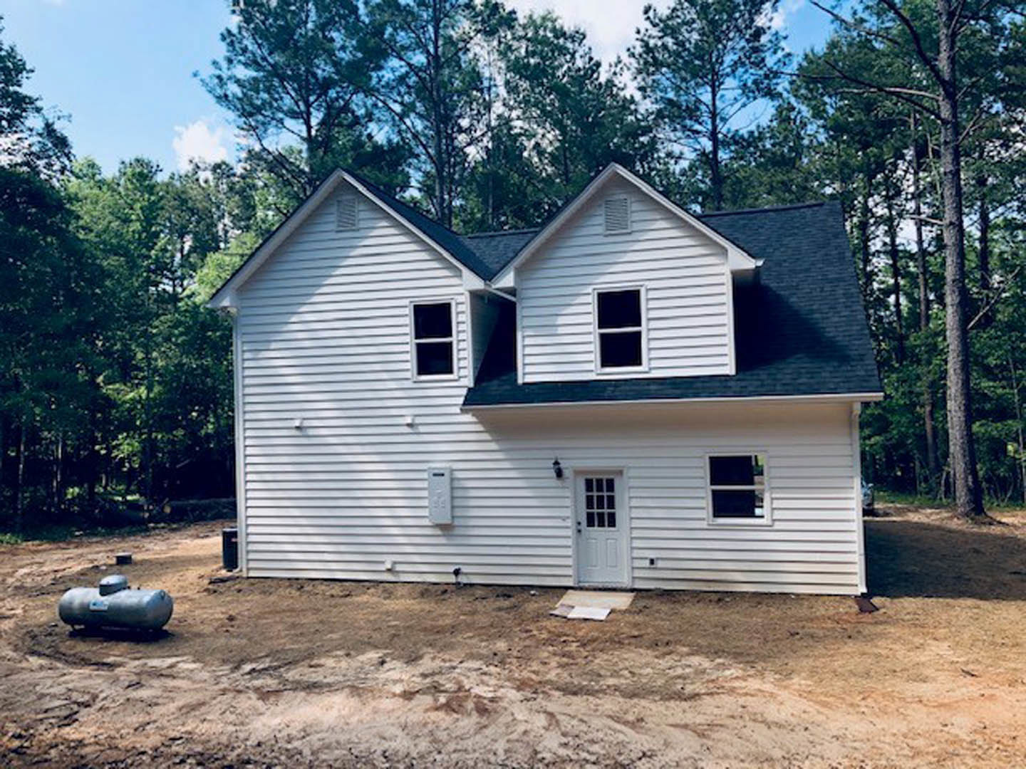 Framed house under construction with exposed wood beams, surrounded by trees and dirt ground, white door with glass panes leaning nearby