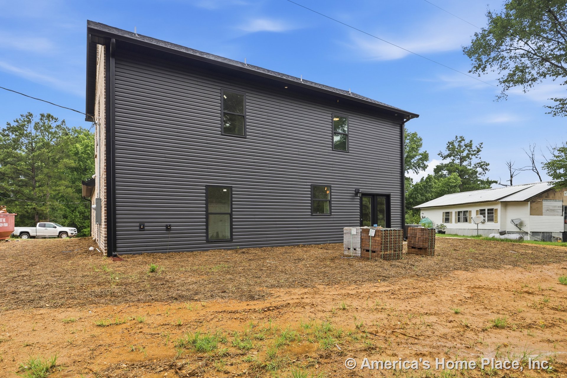 Dark horizontal vinyl siding with black frame rectangular windows, rear exterior door, modern trim, two-story structure, visible utility connections, unfinished yard.