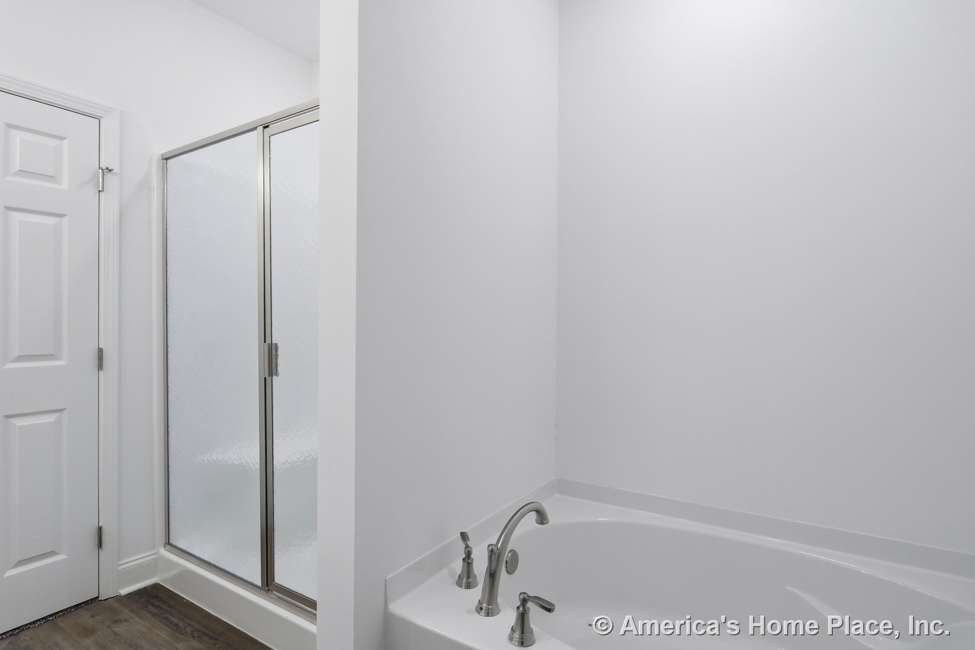 Built-in soaking tub with chrome fixtures beside a glass-enclosed shower, white paneled door, minimalist white walls, wood-look flooring, and simple baseboard trim.