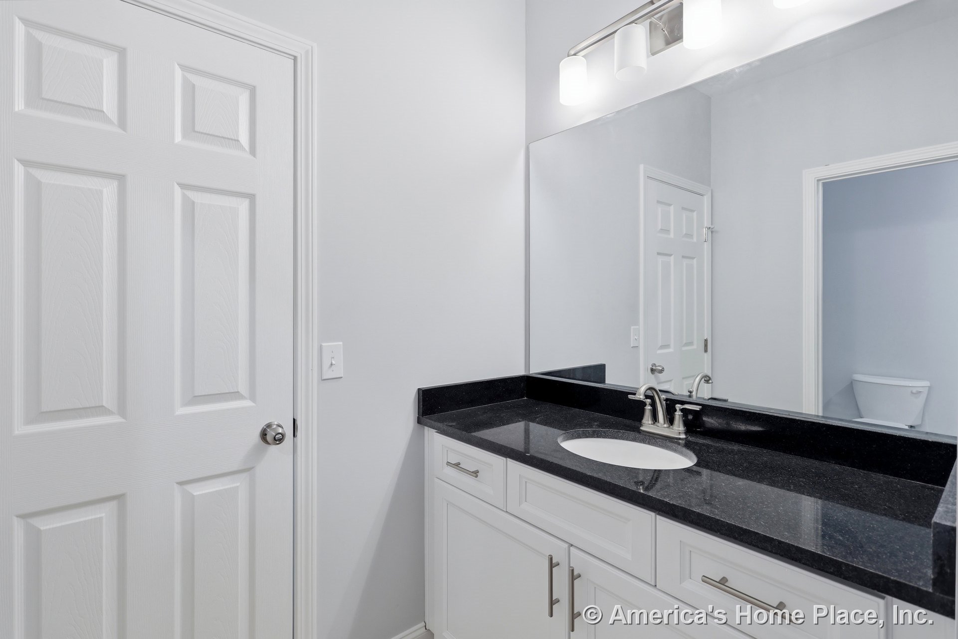 Black solid surface vanity top with undermount sink, brushed nickel faucet, white paneled cabinetry, large wall-mounted mirror, three-light brushed nickel vanity fixture, and white